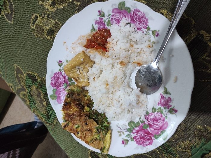A white plate with a floral design sits on a green and gold tablecloth. On the plate are a serving of white rice, a piece of fried tofu, a slice of omelet with green vegetables, and a small amount of red chili sauce. A metal spoon lies on the plate to the right of the rice. The scene is located in the city of Kecamatan Lawang, Indonesia, at coordinates (latitude: -7.828, longitude: 112.701). There are no people in the image.