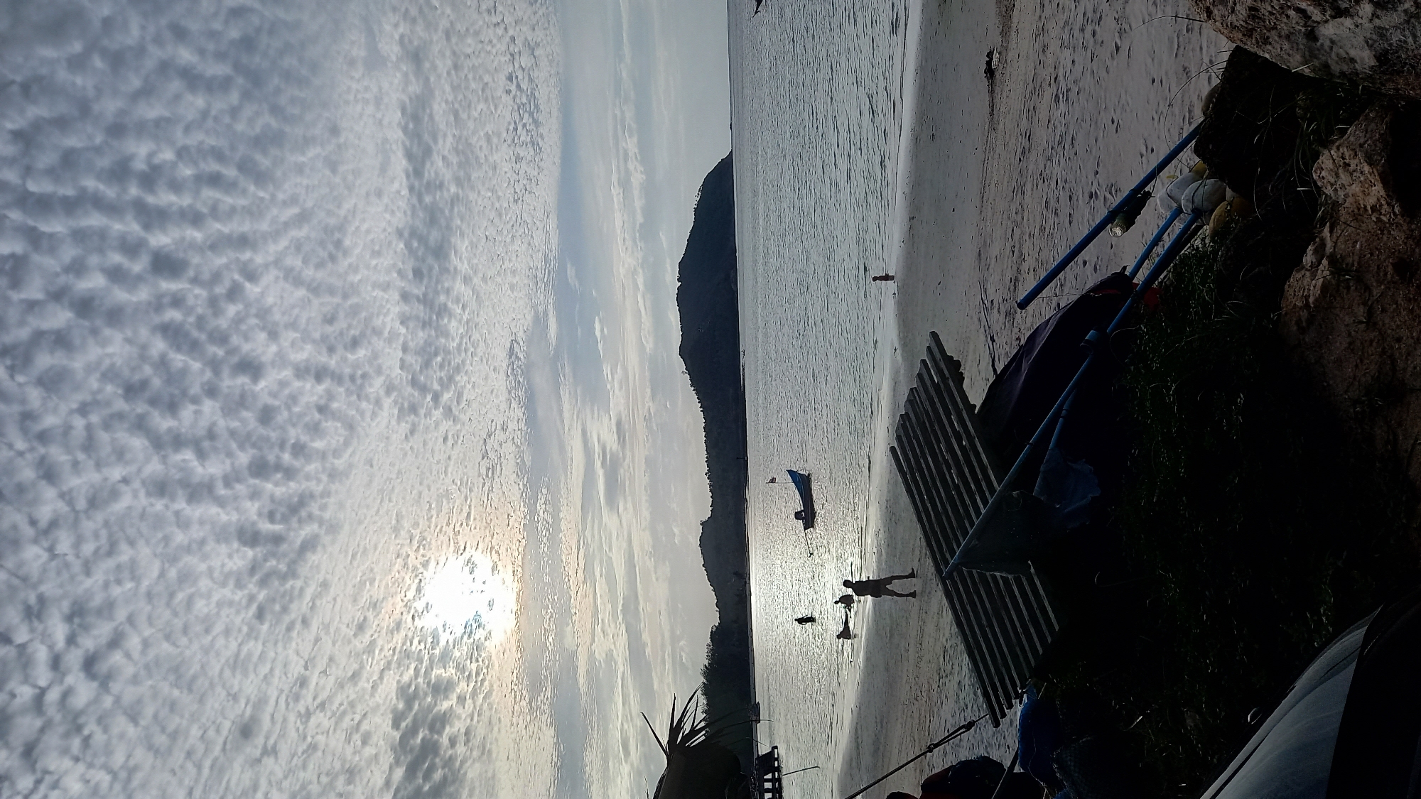 This media file captures a serene beach scene at Koh Phangan, Thailand. The image is oriented vertically, with the sky occupying the left half and the beach and ocean on the right. The sky is filled with a dramatic display of altocumulus clouds, creating a textured, wavy pattern. The sun is partially visible through the clouds, casting a soft, diffused light, suggesting it is either sunrise or sunset.
On the beach, several figures are present. Two individuals, silhouetted against the water, appear to be walking or playing near the shoreline. Another small figure is visible further out in the water. A small boat with a blue sail is anchored in the shallow water. In the foreground, on the right side of the frame, are various objects, including some dark fabric, what appear to be fishing poles or supports, and rocks. Some green vegetation is also visible. The overall mood of the scene is peaceful and tranquil, evoking a sense of calm. The location context indicates this is likely a scene from the coast of Koh Phangan, Thailand. No visible text is present in the image.