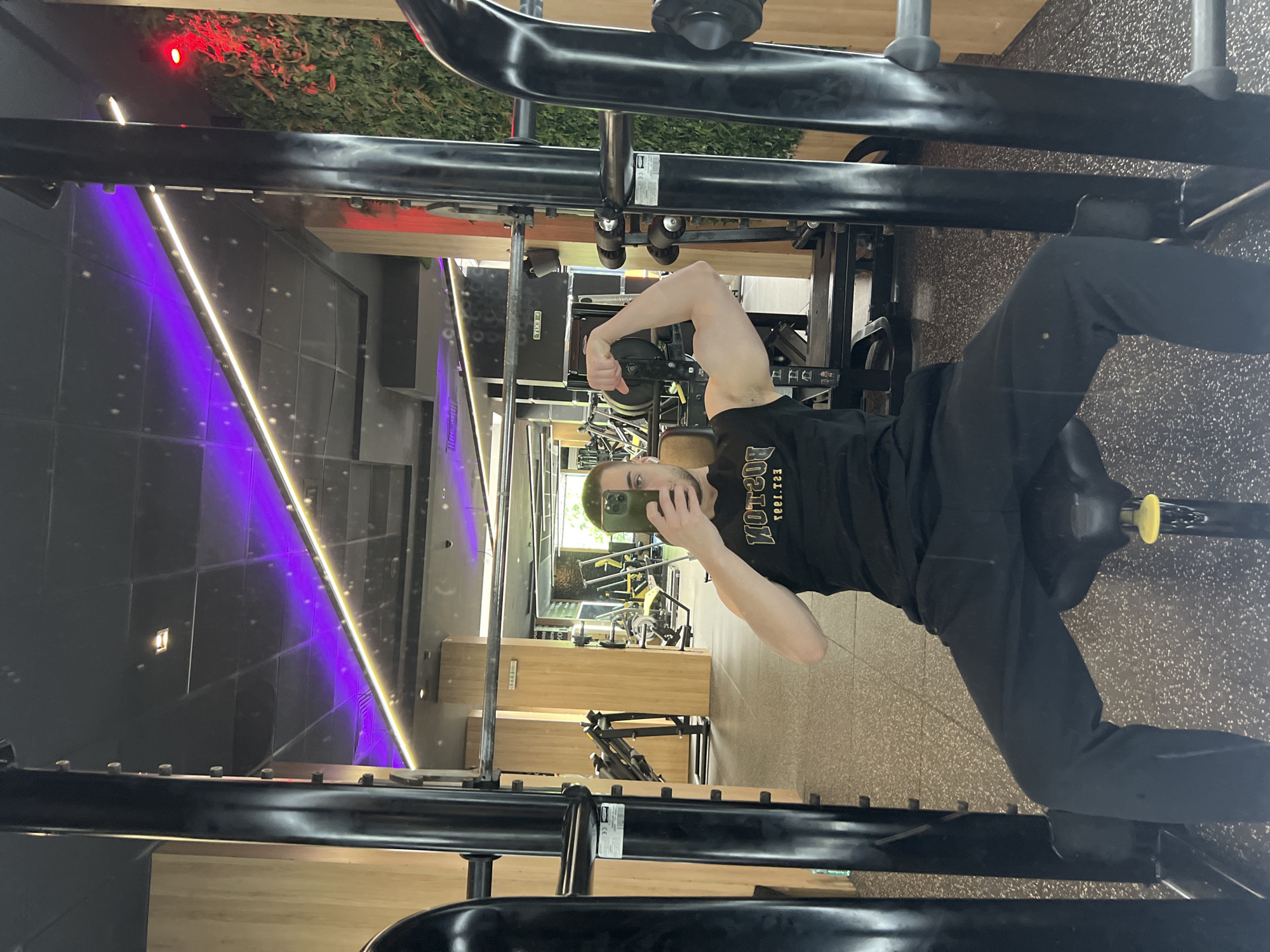 A young adult male is captured in a mirror selfie inside a modern gymnasium in Burgas, Bulgaria. He is seated on a black gym bench, part of a smith machine, and is flexing his left bicep while holding a green-cased smartphone to capture his reflection. He wears a black tank top with a reversed gold-colored logo that reads "BOSTON EST. 1983" and black athletic pants. White earbuds are visible in his ears. The gym features a speckled dark gray rubber floor, light wooden wall paneling, and a prominent artificial green wall accented by a red light on the right. The ceiling is black with grid panels, linear purple and white LED light strips, and recessed lighting. In the reflection behind the subject, additional gym equipment, including weight racks and various machines, is visible. A reversed "EXIT" sign is also discernible on a wall in the background reflection.