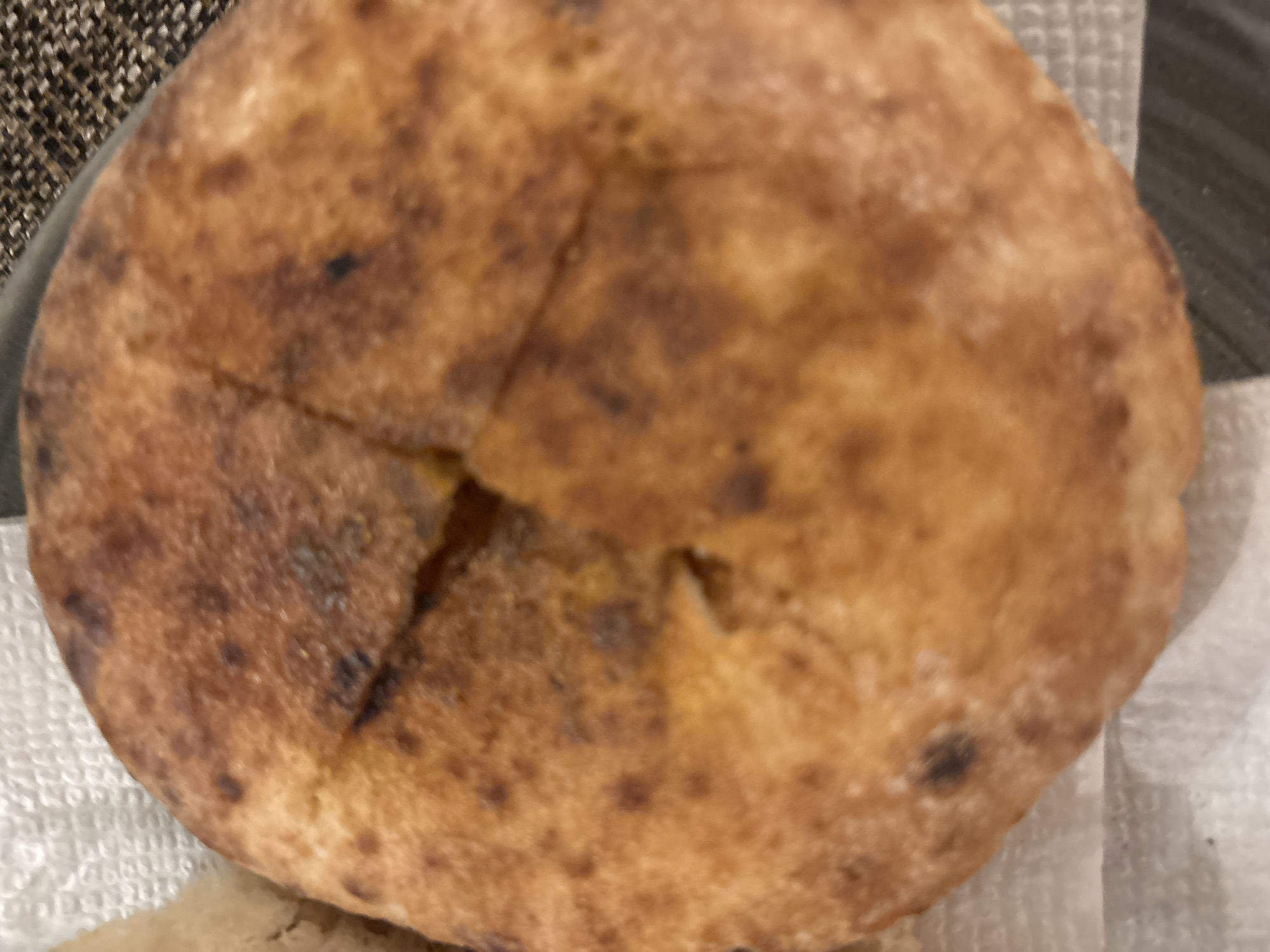 A close-up image shows a round, baked flatbread, possibly pita or similar, with a mottled golden-brown crust. Four diagonal cuts are visible in the center of the bread. The bread sits on a white paper towel or napkin, which is partially visible. The background includes a dark surface and a textured, patterned material. The location and description of Sofia, Bulgaria, are not evident in this image.