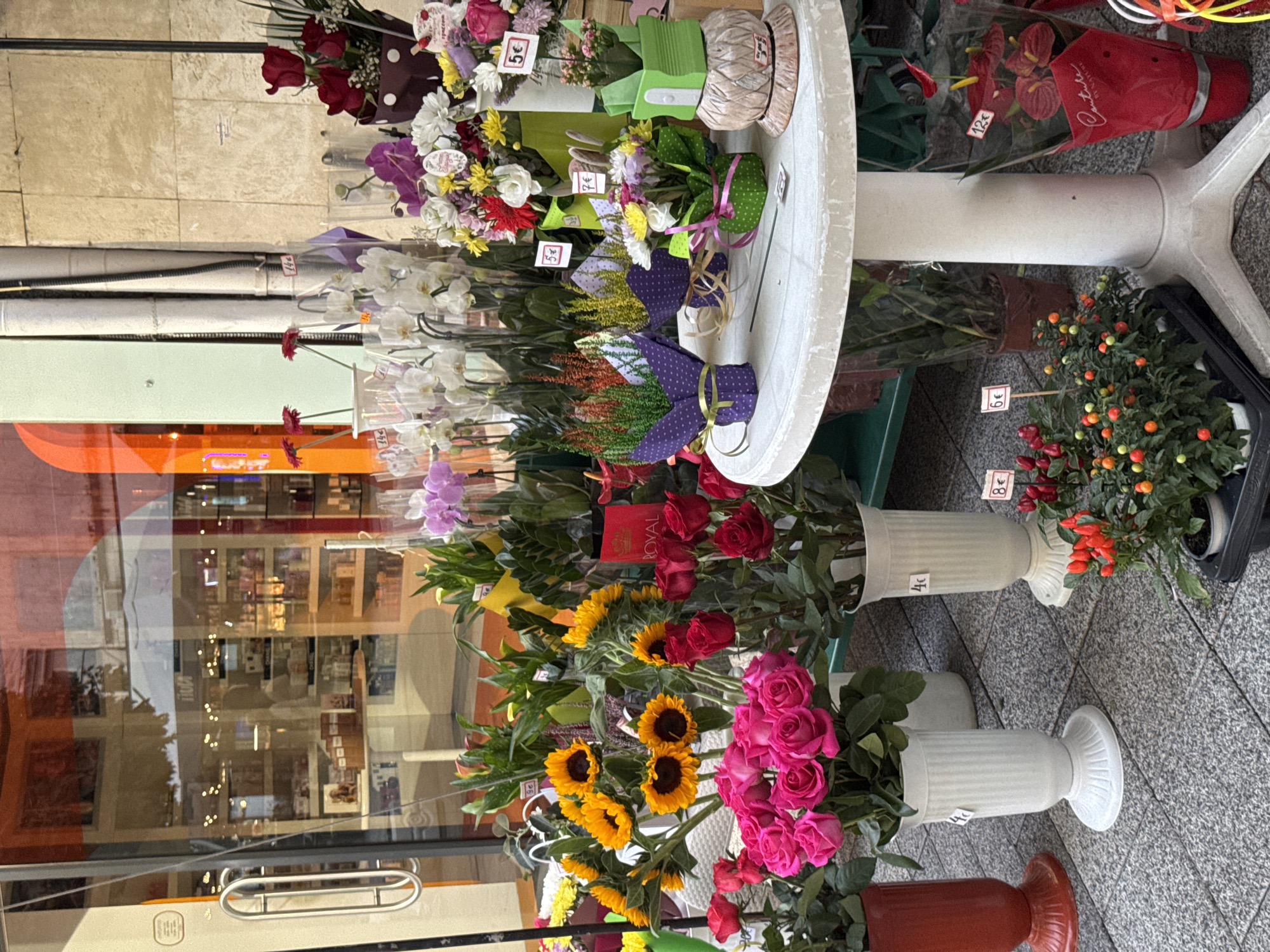 In Plovdiv, Bulgaria, a florist shop's exterior features a display of fresh flowers and potted plants on a sidewalk. In the foreground, a round white table holds several small potted plants wrapped in decorative paper and price tags indicating prices in Euros. To the left of the table, a collection of bright pink roses is displayed in a white vase. Behind the table, a variety of sunflowers, red roses, and orchids are arranged in clear and colored vases, with some plants visible inside the shop through the glass window. The shop's interior shows shelves stocked with products, suggesting it sells cosmetics or fragrances. The ground is paved with gray stone tiles. The overall scene captures a vibrant street-level flower display, typical of urban commercial areas.