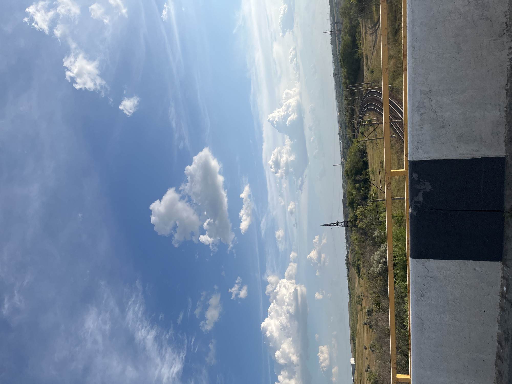 This is a photograph taken from an elevated position, likely a bridge or overpass, looking out over a rural landscape under a bright, cloudy sky. In the foreground, a concrete barrier painted with a black and white stripe is visible, topped with a yellow railing. Beyond the barrier, a grassy field stretches out, dotted with trees and shrubs. A set of train tracks curves through the landscape, disappearing into the distance. Several utility poles and a tall metal tower are also present. The sky is a vibrant blue, filled with scattered white clouds. The location is identified as Ferma, Russia. There are no people or animals visible in the image. The scene appears to be tranquil and undeveloped.