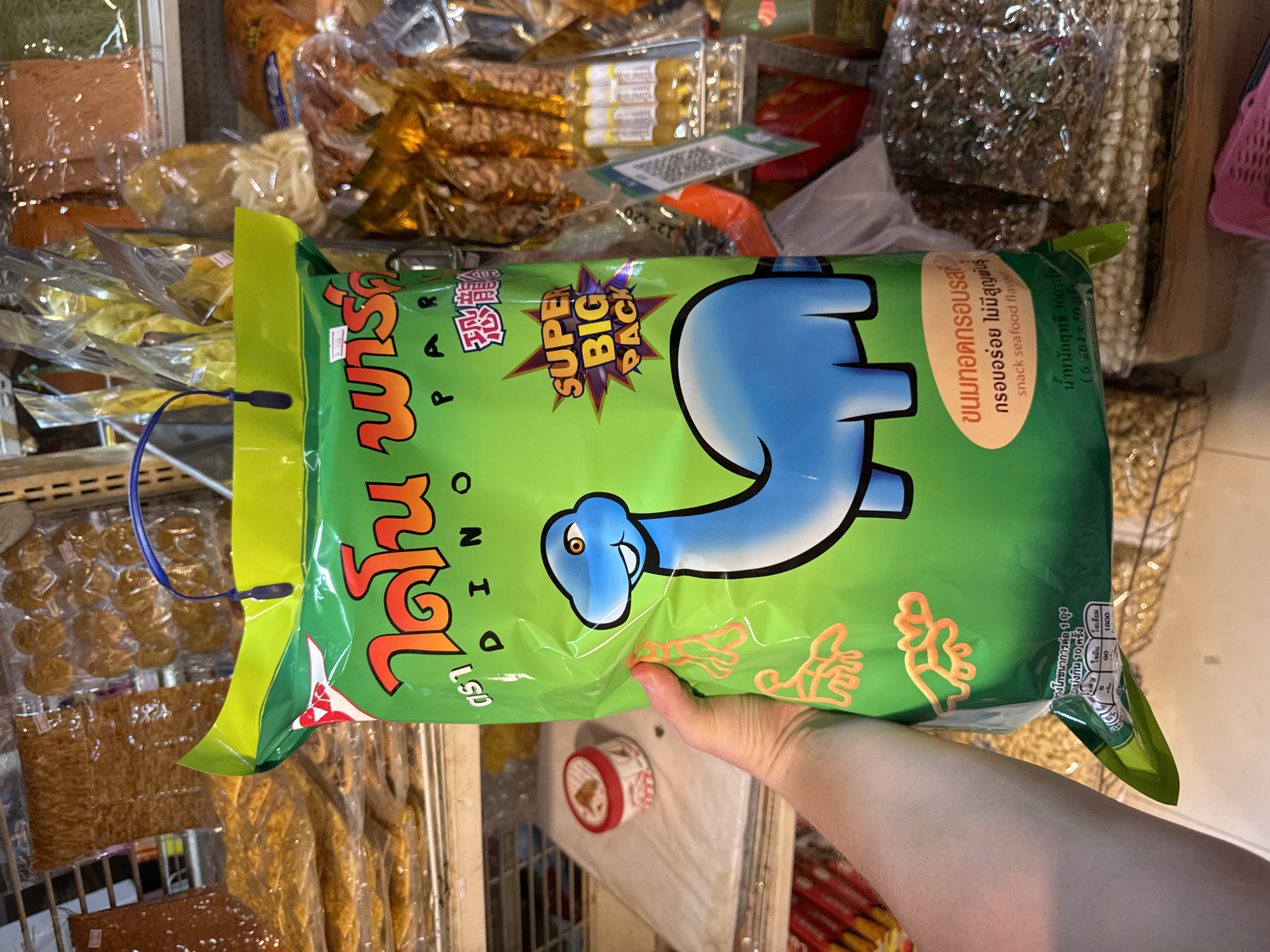 An indoor, slightly overhead shot captures a person's hand holding a large, bright green snack bag, likely within a market or grocery store aisle in Pattaya City, Thailand. Various other packaged food items, including dried goods and snacks, are visible in the blurred background, stacked on shelves and in bags.
The prominent snack bag is decorated with a friendly blue dinosaur character. Thai script on the bag reads "DINO" and includes the Chinese character for "dinosaur" (恐龍). A starburst graphic highlights "SUPER BIG PACK." Additional Thai text identifies the product as a "crispy fried snack" with a "delicious, never extinct" tagline and specifies it is a "snack seafood flavor." The net weight is indicated as "300 grams (6 sachets x 50 grams)." Nutritional information states "Nutrition per 1 bag" and suggests "10 servings," listing values for fat (90) and sodium (1800).
The person's hand, with a light skin tone, holds the bottom right corner of the bag. A blue cord, possibly from headphones or a price scanner, is visible near the top left of the bag. The overall lighting is bright and consistent with an indoor retail environment.