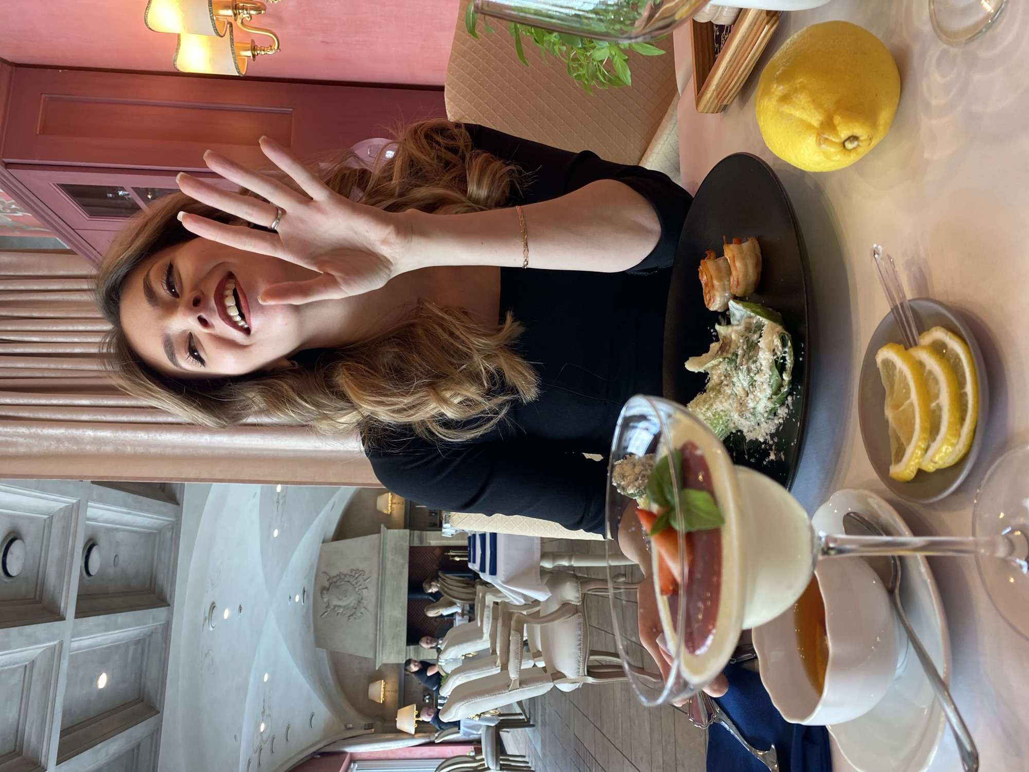 A smiling woman with long brown hair, wearing a black top, is seated at a table in a restaurant. She is looking towards the camera and holding her right hand up, revealing a silver ring on her ring finger. A delicate gold bracelet is visible on her left wrist.
In front of her, on the light-colored tablecloth, is a black oval plate containing what appears to be a salad (possibly Caesar salad) topped with grated cheese, and a few cooked shrimp or prawns. To the right of the plate, a small grey dish holds several lemon slices. A whole, rough-skinned lemon rests beside it. Further to the right, a martini glass holds a dessert, possibly a panna cotta, garnished with red fruit (strawberries) and a green leaf (basil). This glass sits on a white saucer with a spoon and a white cup. Another glass, partially visible behind the woman's arm, contains a green plant, likely herbs.
The restaurant features an elegant interior with high, arched ceilings, decorative plasterwork, and recessed lighting. In the background, other patrons are seated at tables with white upholstered chairs. A large, ornate fireplace-like structure with carved details is visible. Warm-toned chandeliers hang from a lower ceiling section near the foreground. The overall atmosphere suggests an upscale dining experience. The scene takes place in a restaurant in Yekaterinburg, Russia. There is no visible text in the image.