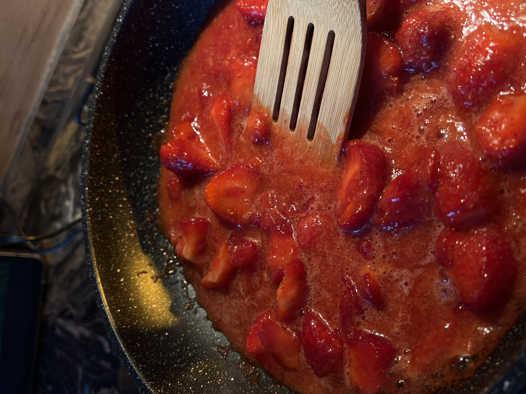 The image displays a close-up, high-angle view of a black non-stick frying pan filled with cooking strawberries. Large, cut pieces of bright red strawberries are submerged in a bubbling, deep red liquid, indicating they are being cooked down, possibly for a sauce or jam. Numerous small bubbles are visible across the surface of the simmering mixture. A light-colored wooden slotted spatula, featuring three horizontal slots, is partially visible on the right side, resting within the strawberry mixture. The interior edge of the pan shows a bright golden-orange reflection on its black surface, suggesting an overhead light source. The background is out of focus, revealing dark, indistinct kitchen elements, including what appears to be a countertop and blurred appliance components in the upper area. No individuals are present in the scene. The depicted location is the city of Ternopil, Ukraine.