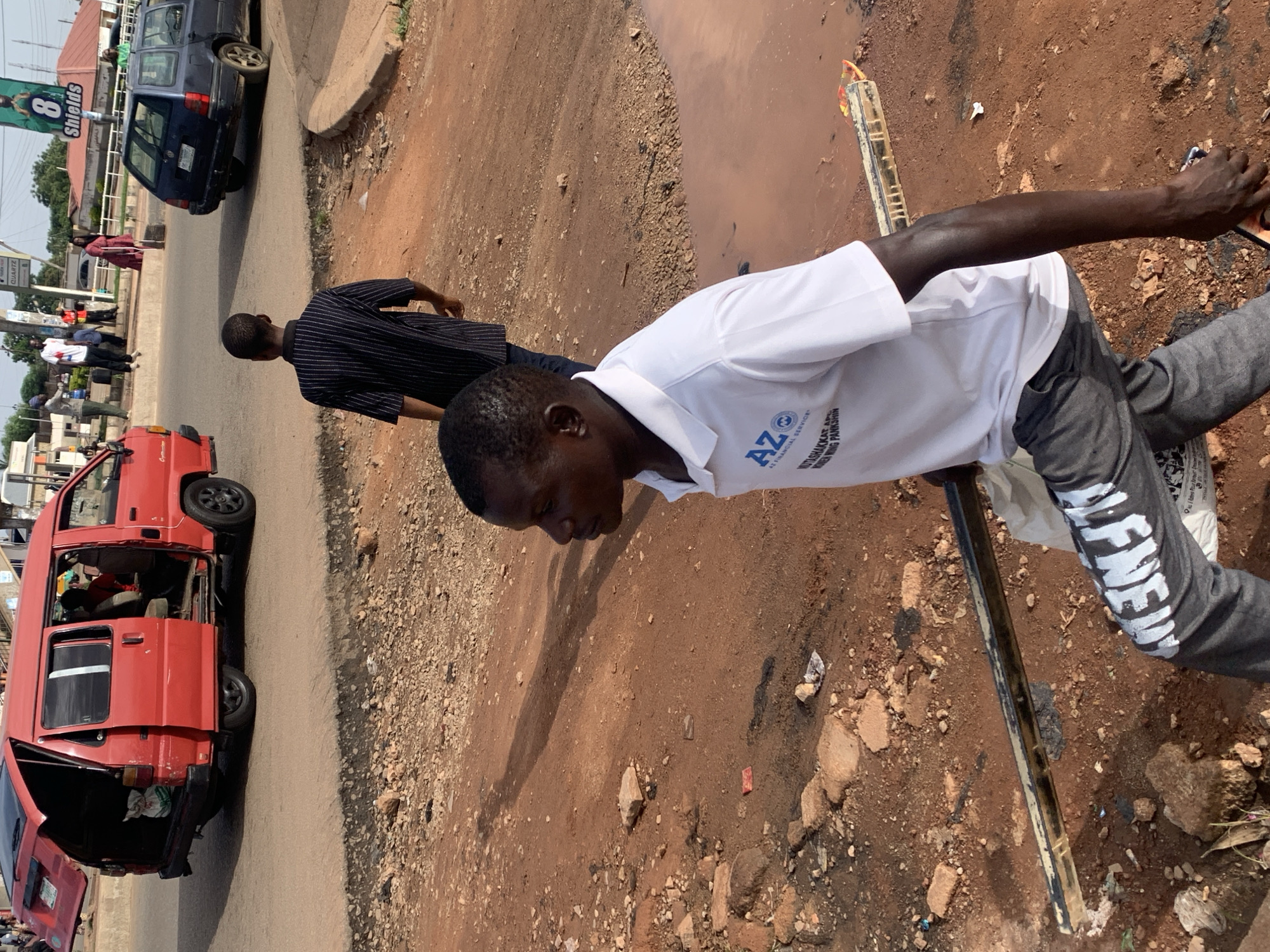 The image depicts a street scene in Gura Top, Nigeria, on a sunny day. In the foreground, a Black man wearing a white polo shirt with an "AZ FINANCIAL SERVICE" logo and dark grey pants with white lettering is walking towards the right, carrying a long, dark-colored object. His gaze is directed ahead and slightly to the right. Behind him and to the left, another Black man in a striped shirt is walking away from the camera.
To the left of the frame, a red minivan with its rear door and passenger door open is parked on the side of the road. Several people are visible inside and around the minivan. Further down the road, a dark blue station wagon is driving away from the camera. The environment consists of a dirt road with scattered rocks and debris, and a large puddle of reddish-brown water on the right side of the image. A billboard with the number "8" and the word "SHIELDS" is visible in the background, along with buildings and other vehicles. The overall impression is of a busy, dusty street in an urban or semi-urban area.