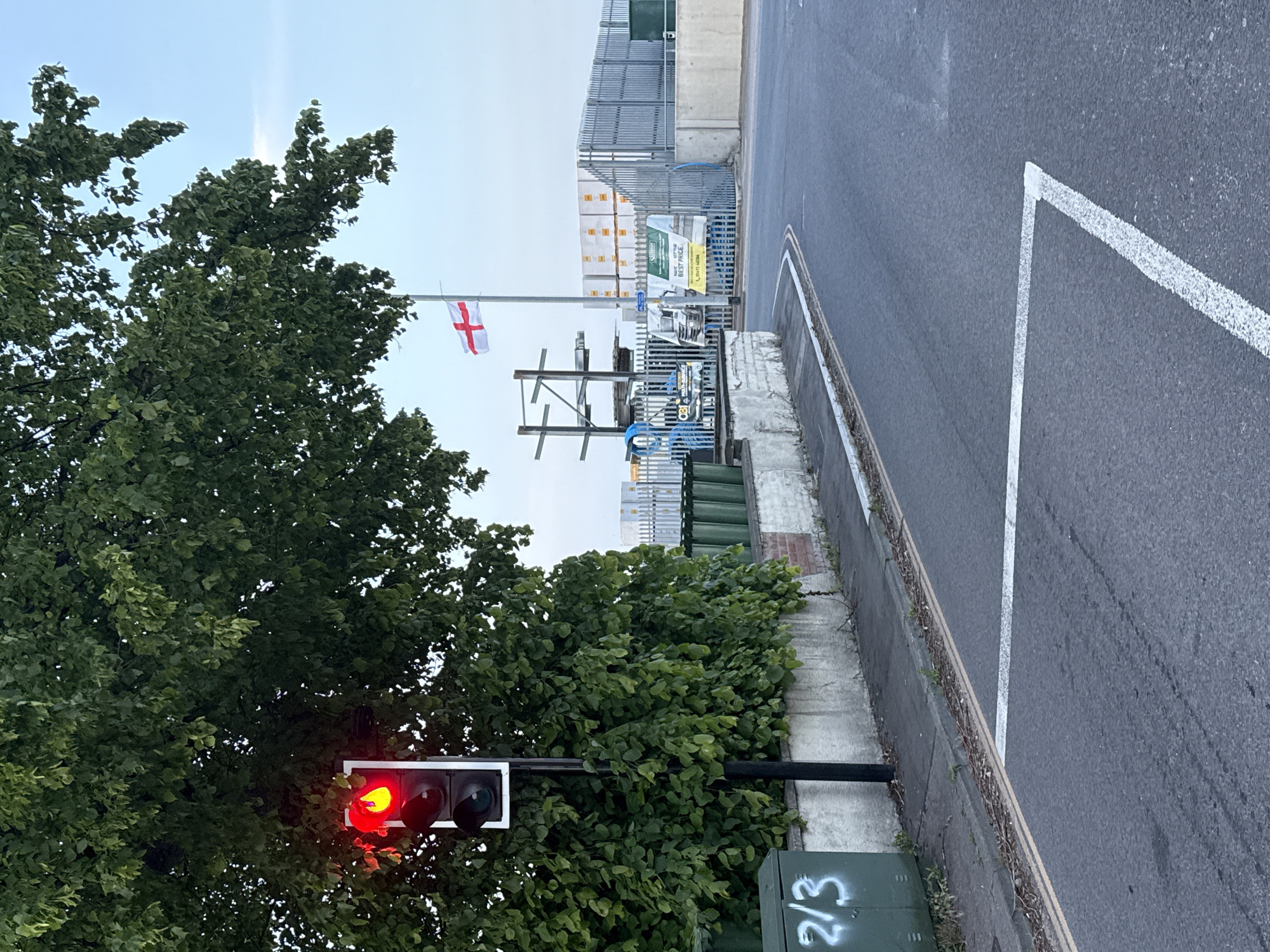 The image depicts a street scene with a red traffic light, partially obscured by a tree, and a flag of England flying from a pole in the background. The road is asphalt with white painted lines marking a pedestrian crossing. A metal fence and stacked building materials are visible behind the flagpole. A concrete wall runs alongside the road, and a green electrical box with white graffiti is located near the traffic light. The scene appears to be in Great Yarmouth, United Kingdom based on the English flag.