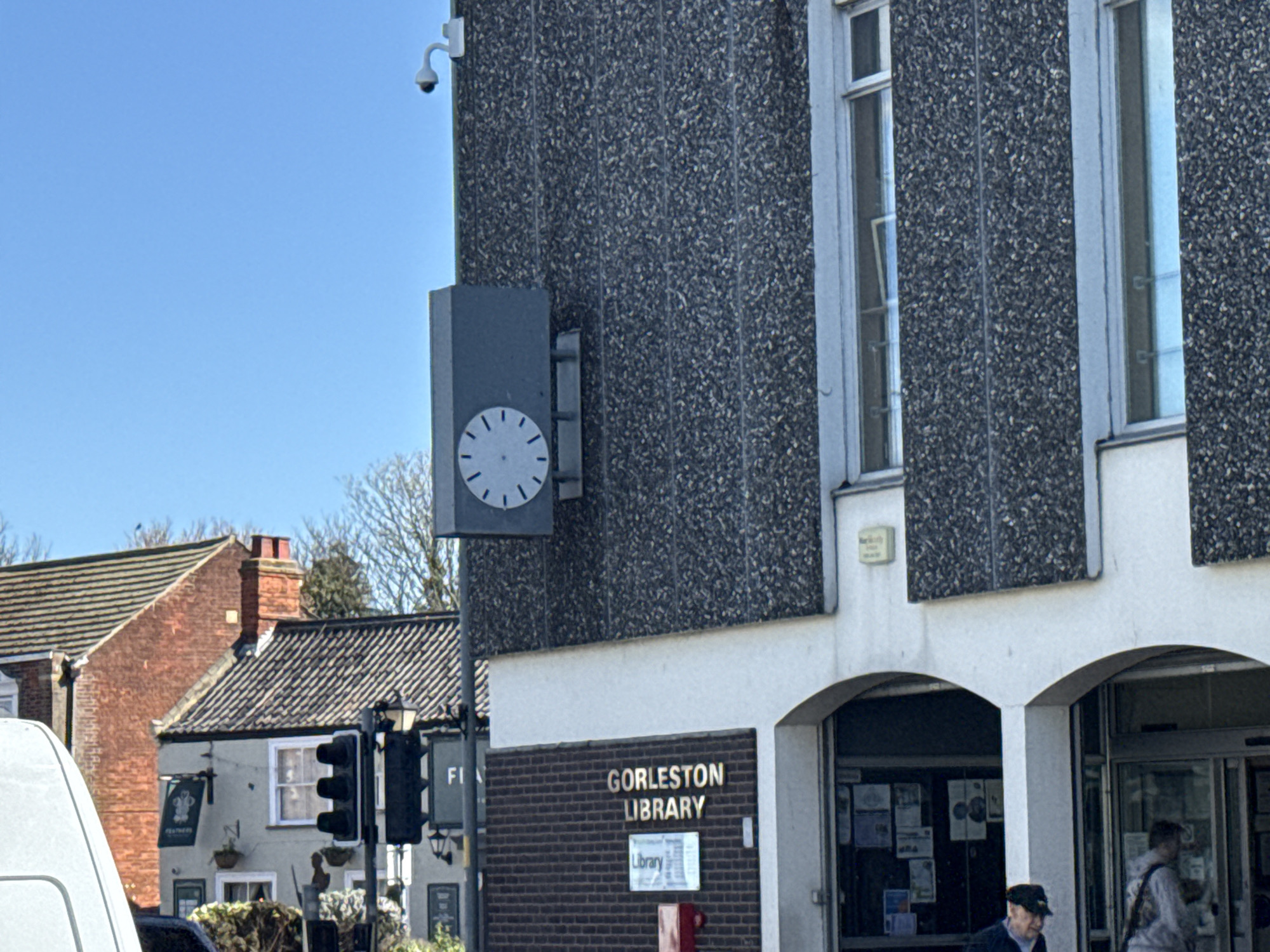 The image shows the exterior of Gorleston Library in Gorleston-on-Sea, United Kingdom. The library building is constructed with a textured grey facade and a white base. A clock is mounted on the building's exterior, and a security camera is positioned above it. Signage for the library is visible on the brickwork at the front. In the background, there is a building with a red brick chimney and a pub sign. Pedestrians are entering the library. Traffic signals are also visible near the pub.