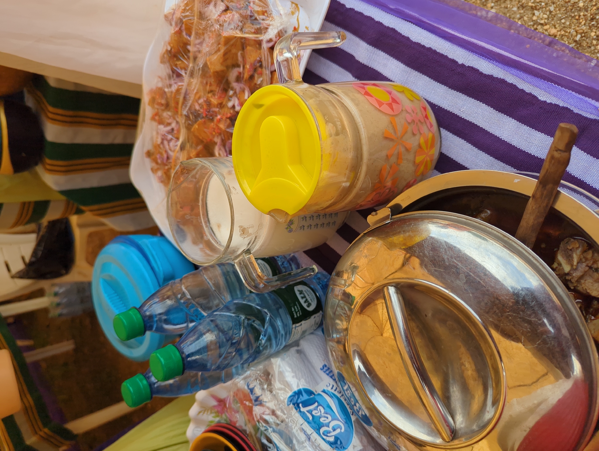 The image presents an outdoor scene with a table setup for a meal, likely in the city of Maigizo, Nigeria. The table is covered with a striped purple and white cloth. Several items are arranged on the table: a clear plastic bag containing fried food mixed with red onion and spices, a glass pitcher with a yellow lid and floral decorations, a clear glass cup, a blue water bottle with a green cap, a pack of "Best" brand serviettes, a stainless steel serving dish with a lid, and a wooden spoon inside the dish. The setting appears casual and utilitarian.