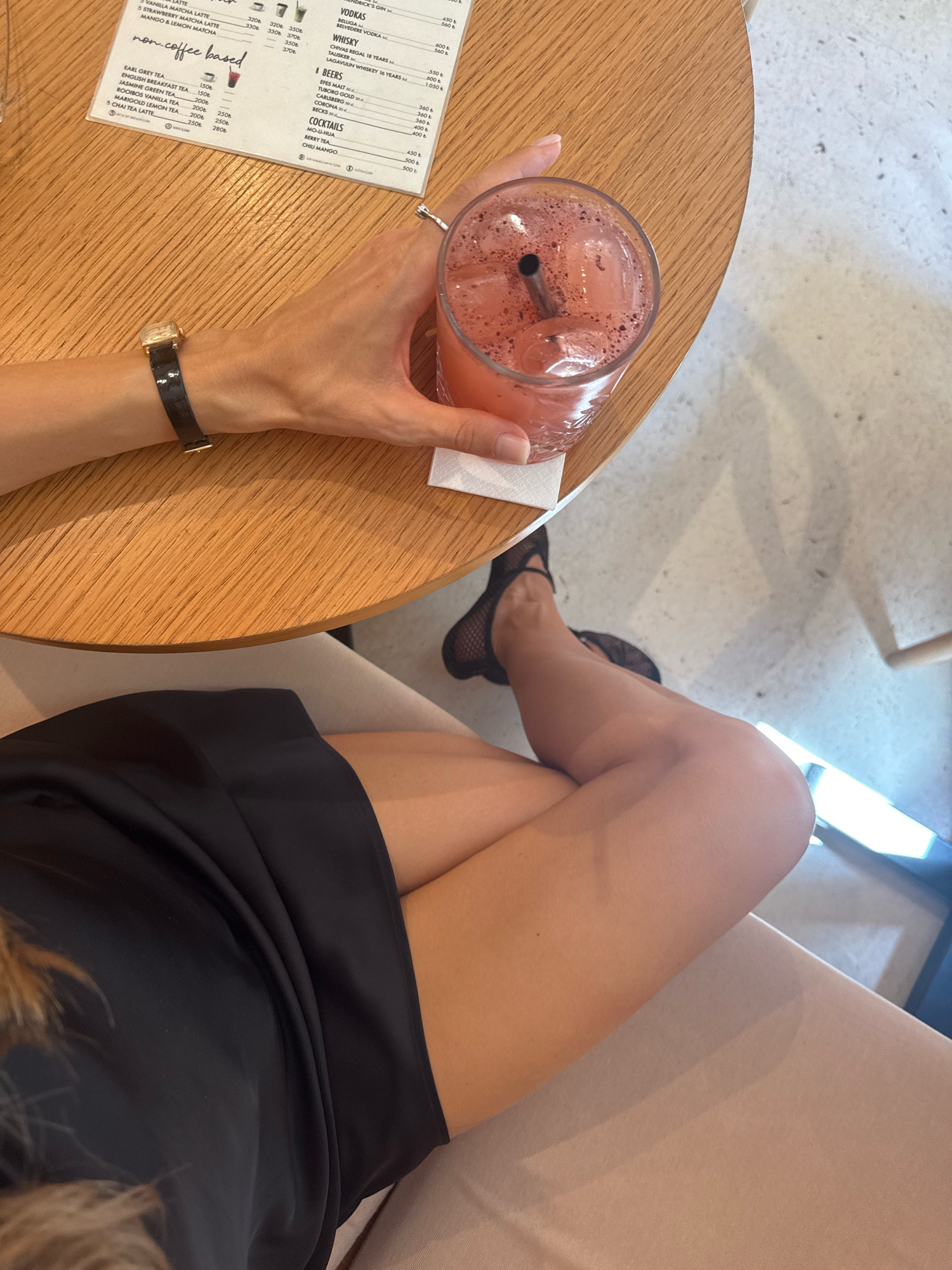 A woman's arm, adorned with a black watch, holds a glass of pink liquid with ice and a dark straw. She is seated at a round, light-wood table. To her right, a menu with various beverage options is visible. The woman is wearing a black dress and black patterned shoes. The scene appears to be indoors, possibly in a cafe or restaurant, with a textured white floor. The lighting suggests a bright, daytime setting. The overall atmosphere is casual and relaxed. The provided crops confirm the presence of a menu listing drinks such as Vodkas, Whiskeys, Beers, and Cocktails. The location is identified as Antalya, Turkey.