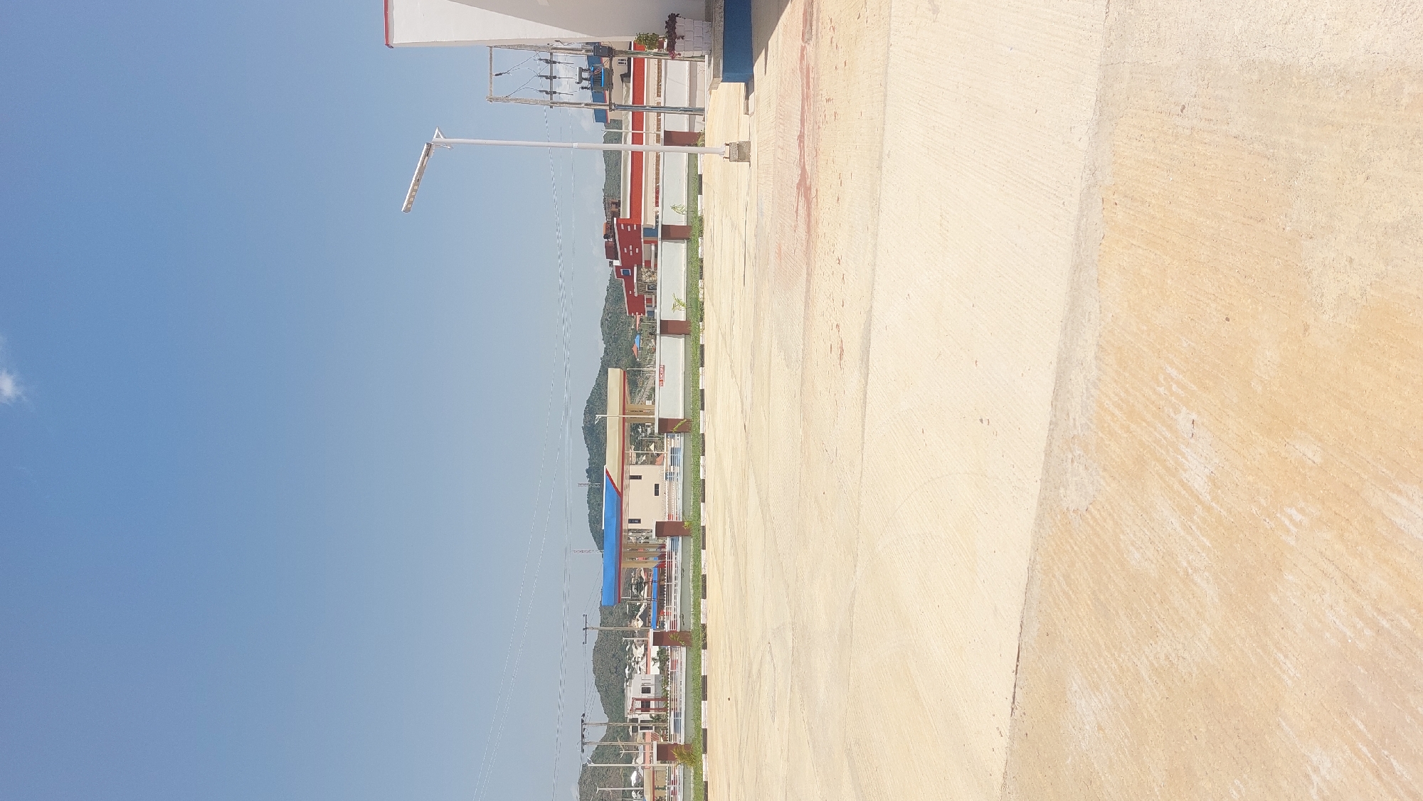 The image displays a bright, sunny day in Kuru Dungus, Nigeria. The scene is dominated by a large expanse of light-colored, textured concrete in the foreground, suggesting an outdoor paved area like a walkway, plaza, or open space. To the left, a row of colorful buildings, likely shops or offices, lines a street. These buildings feature a mix of white, red, and blue elements, with some appearing to be multiple stories high. A street lamp stands tall in front of the buildings, and utility poles with wires are visible, indicating a developed area. In the background, lush green hills rise, with more buildings scattered at their base and along the slopes, hinting at a town or city nestled within the landscape. The sky is a clear, vibrant blue, with a small wisp of a cloud. The overall impression is one of a well-lit, active urban or semi-urban environment on a pleasant day. No people are visible in the frame. There is no discernible text in the image.
