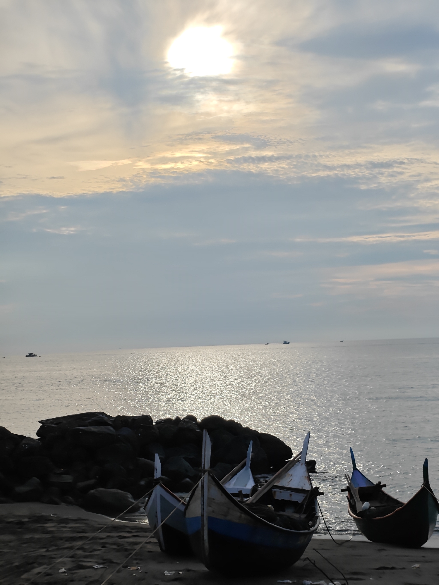 The image depicts a serene coastal scene at dusk in Lhokseumawe, Indonesia. The sun, partially obscured by wispy clouds, casts a shimmering reflection on the calm ocean surface. In the foreground, several traditional fishing boats are beached on the sandy shore. The boats are of varying sizes and colors, with some appearing worn and weathered. They are tethered to the shore with ropes. Dark, rocky outcrops are visible along the coastline, contrasting with the lighter sand. In the distance, a few more boats can be seen on the water, adding to the sense of a working fishing community. The overall atmosphere is peaceful and tranquil, characteristic of a quiet evening by the sea.