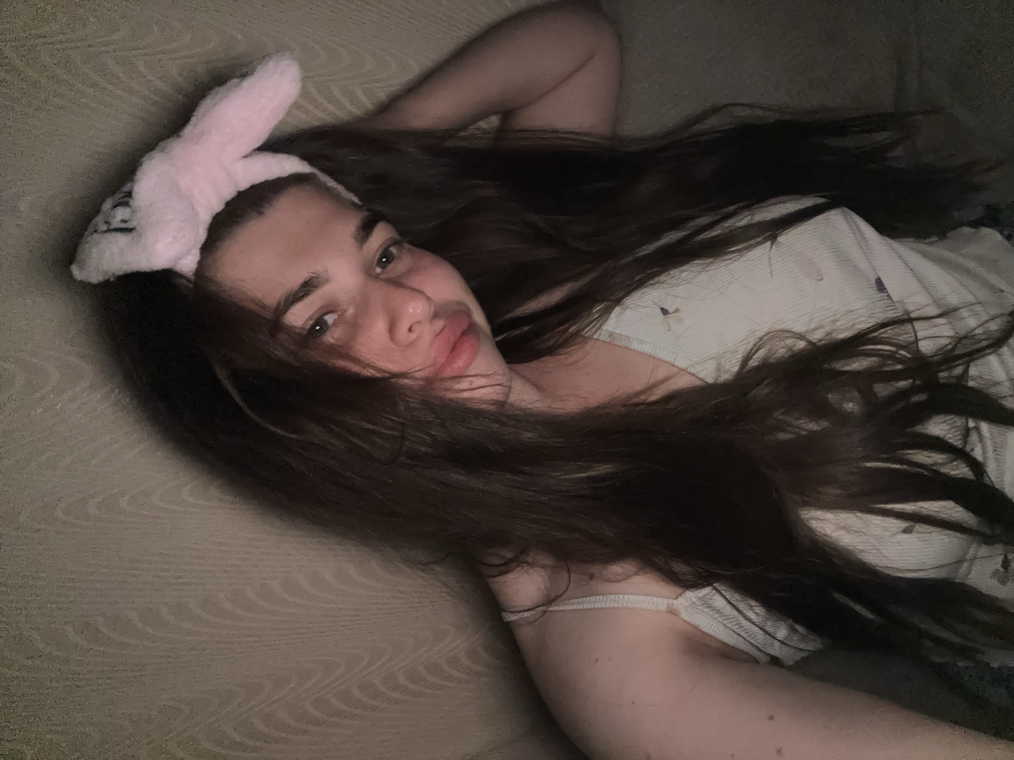 In this selfie taken in the city of Мещёры, Russia, a young person with long, dark brown hair is looking at the camera. They are wearing a light-colored tank top with a delicate pattern and a fluffy pink headband with bunny ears. The background appears to be a textured wall or fabric, possibly a couch or a wall covering, with a wavy pattern. The lighting is dim, creating shadows and highlighting the person's face and hair. The person's left hand is resting near their face, and their right arm is bent at the elbow, with their hand near their head. The overall impression is that of a casual, intimate moment captured in a domestic setting.