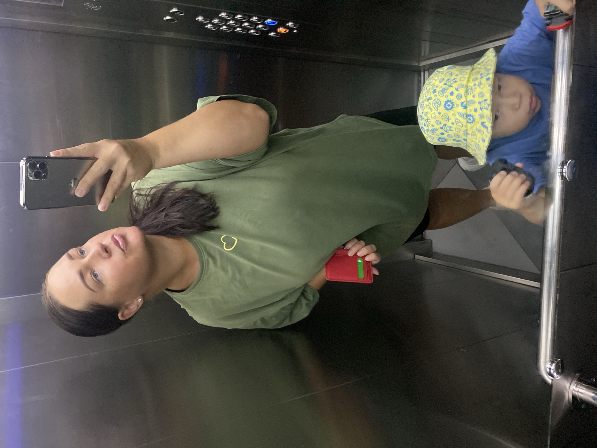 An indoor elevator selfie is taken, reflecting in the mirror. A woman with her hair pulled back, wearing a green t-shirt with a yellow heart graphic, holds a red cardholder in her left hand. She is looking to her left, with her right hand holding a dark-colored smartphone up to take the picture. A young child wearing a yellow bucket hat with a blue pattern and a blue t-shirt is visible in the reflection, leaning on a railing and holding two small black objects. The elevator walls are metallic and reflective, with a panel of buttons visible on the right side. The floor appears to be tiled. The location is identified as an elevator in Sochi, Russia.