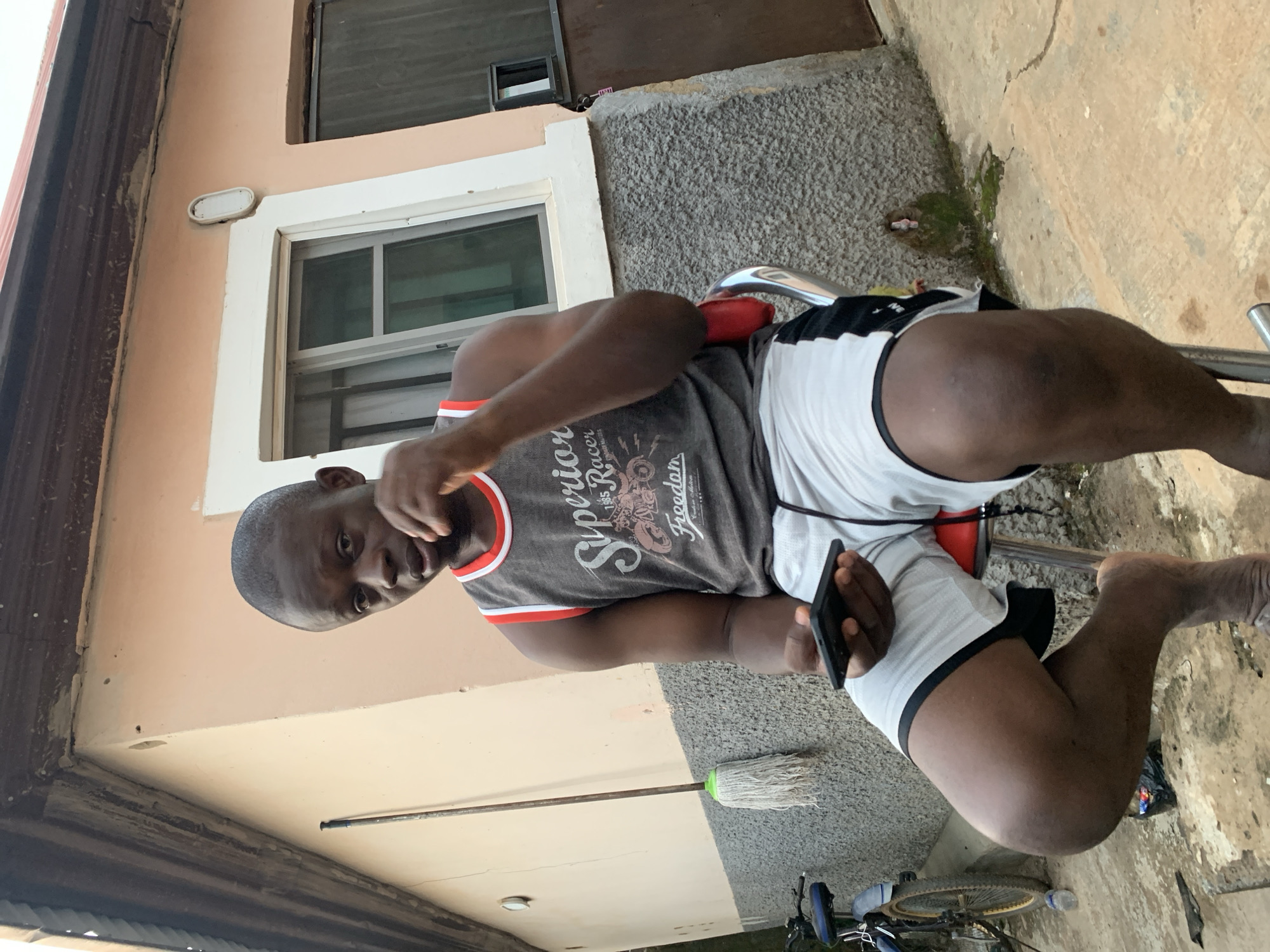 A man is sitting on a red and chrome stool outdoors. He is wearing a grey tank top with red trim, white shorts with black trim, and he is holding a black smartphone in his left hand. His right hand is gesturing towards the camera. Behind him is a beige building with white trim around a window. There is a mop next to his leg and a bicycle on the ground in the background. The location is potentially Abuja, Nigeria, but without further context or evidence, this cannot be definitively confirmed. The background appears to be a private residence or yard, based on the exterior of the building and the presence of personal items such as the mop and bicycle.