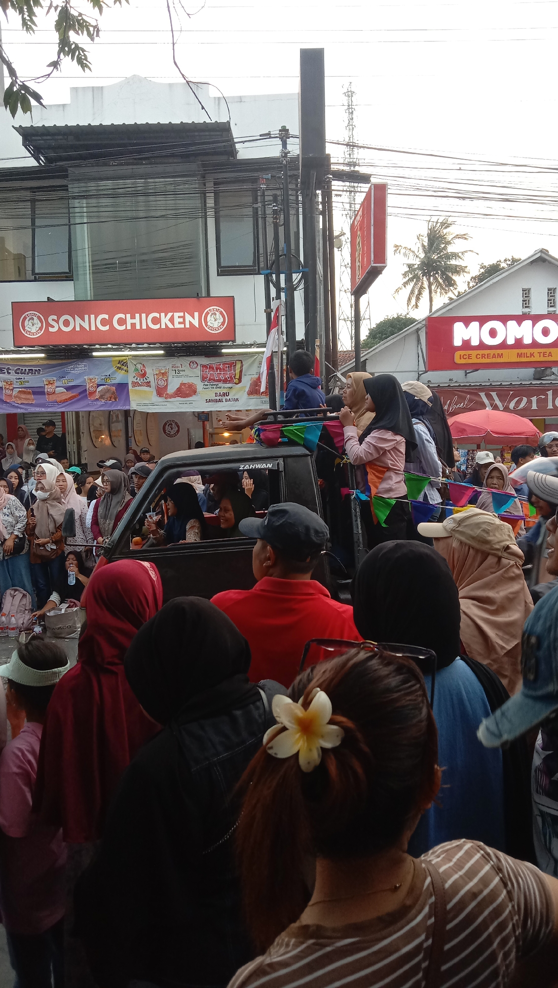 An outdoor street scene in Jatisawit, Indonesia, shows a crowd of people gathered around a black pickup truck. The truck is decorated with colorful triangle flags and has several people riding in the bed and standing on top. Some people in the truck are holding drinks and smiling. The crowd is diverse, with many women wearing hijabs. In the background, there are storefronts with signs for "SONIC CHICKEN" and "MOMO ICE CREAM MILK TEA". A palm tree is visible to the right. The overall impression is one of a festive gathering or event. The lighting suggests it is either early morning or late afternoon.