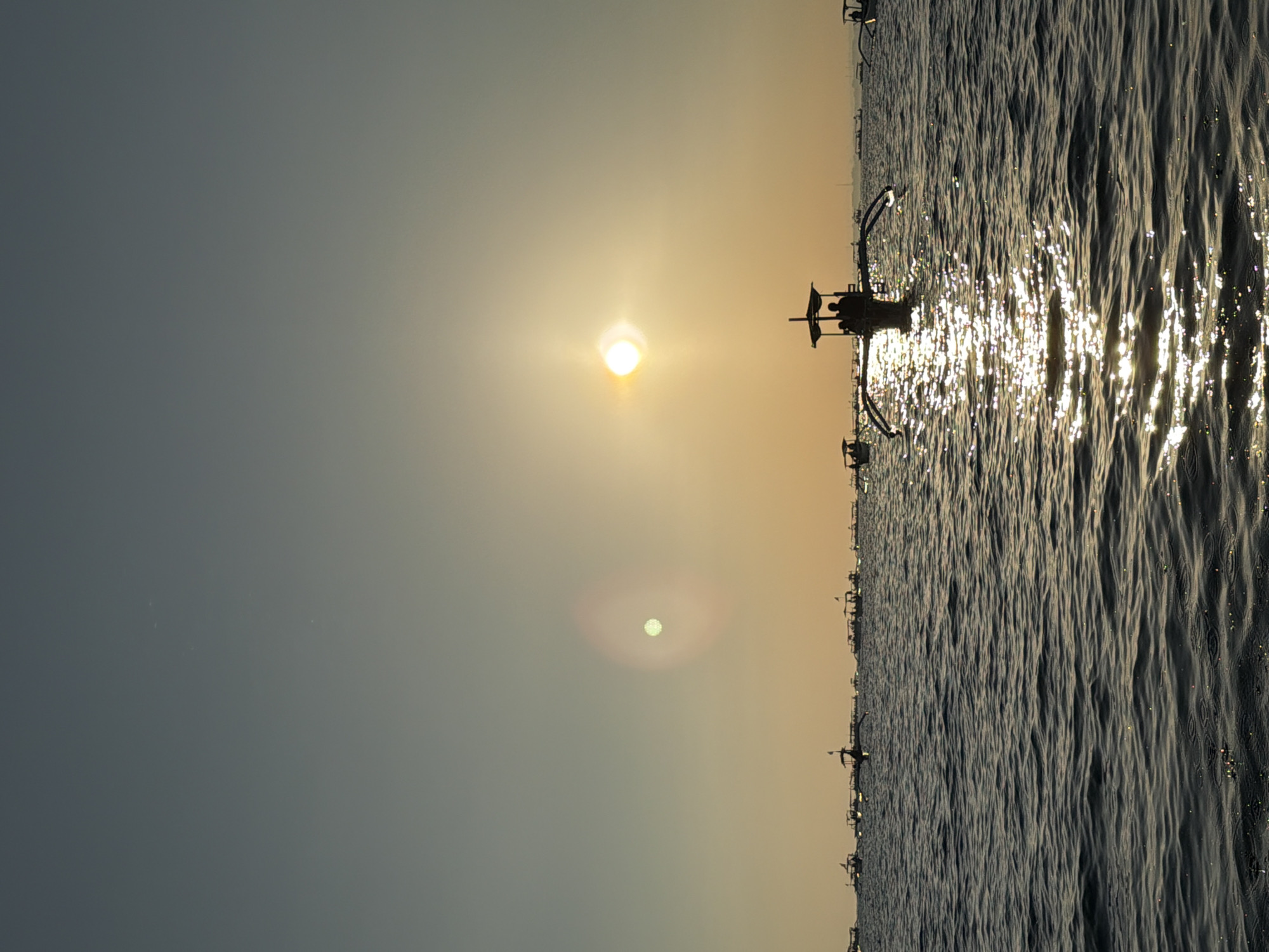 The image depicts a body of water, likely the ocean, under a hazy sky with the sun low on the horizon, suggesting either sunrise or sunset. The sun is a bright, diffused orb casting a warm glow and creating a reflective shimmer on the water's surface. In the foreground, the water exhibits gentle ripples, with glints of light scattered across it. A line of silhouetted traditional boats, known as outrigger boats, are positioned in the middle ground, stretching across the frame. One prominent boat in the center features two figures standing under a canopy. The background reveals a further scattering of similar boats, also silhouetted against the bright sky. The overall scene evokes a serene and tranquil coastal environment in Indonesia.
