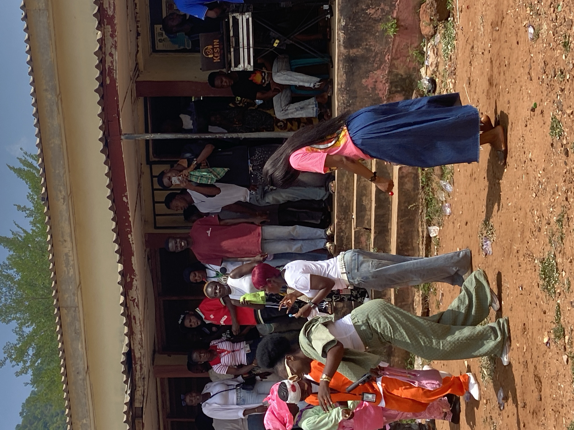 The image shows a group of people gathered outdoors in Lokoja, Nigeria. They are assembled around a building with a corrugated metal roof and faded paint. Some people are standing on the steps of the building, while others are on the ground. A woman with long black hair wearing a pink shirt and blue skirt appears to be performing or dancing in front of the crowd. Some individuals are taking photos or videos with their phones. A sound system labeled "KESIN" is visible in the background. The ground is reddish-brown dirt with scattered debris.