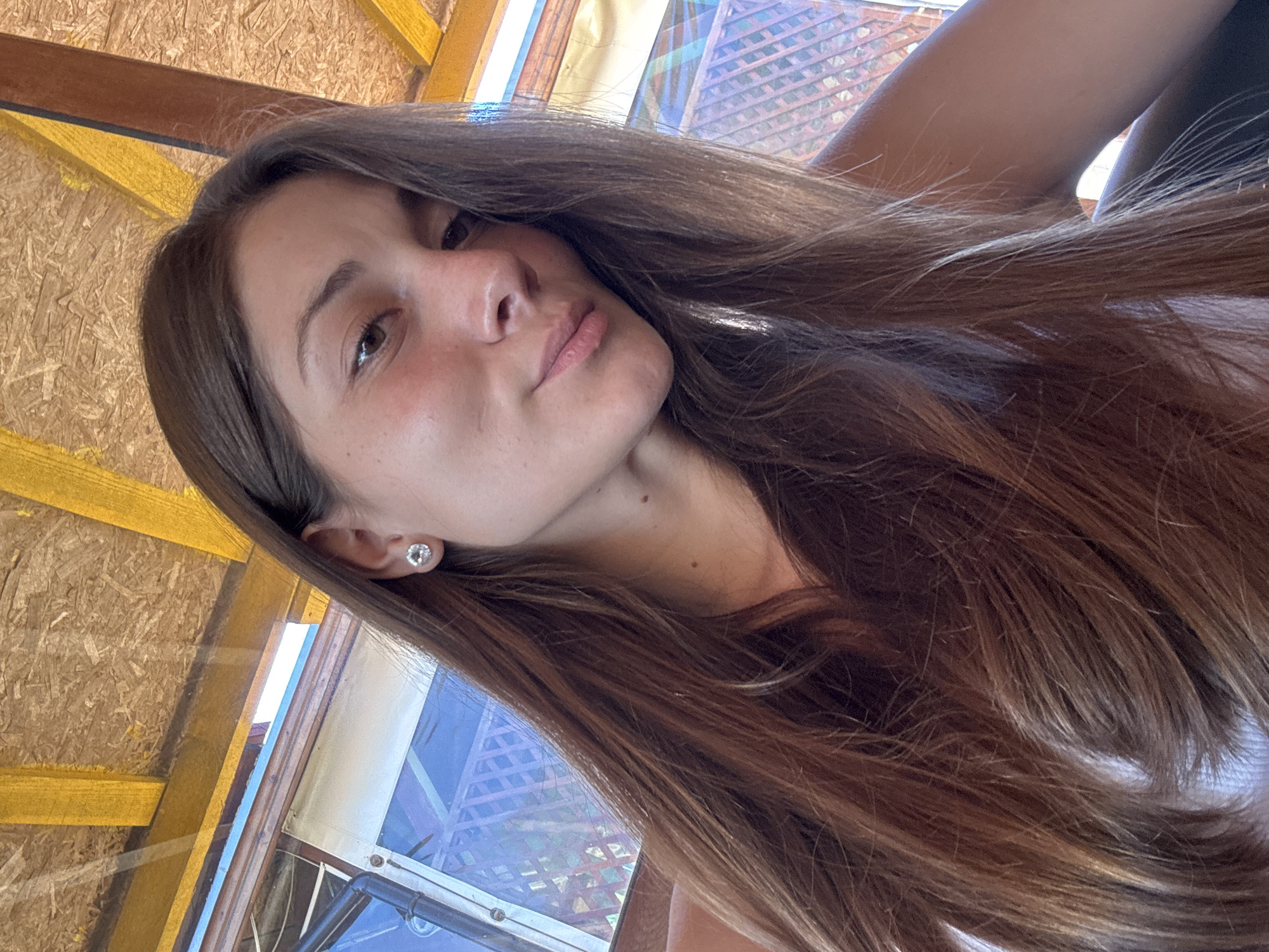 The image is a close-up, low-angle selfie of a young woman with long, brown hair. She is looking towards the camera with a slight smile, her head tilted. She is wearing a white top and a single stud earring in her left ear. The background appears to be an outdoor, possibly covered, area with wooden beams and what looks like particle board ceiling. In the mid-ground, a window or screen with a latticed pattern is visible, suggesting a location that might be a cafe, restaurant, or residential outdoor space. The lighting suggests it is daytime. The context of Sozopol, Bulgaria, is not visually verifiable from the image itself.