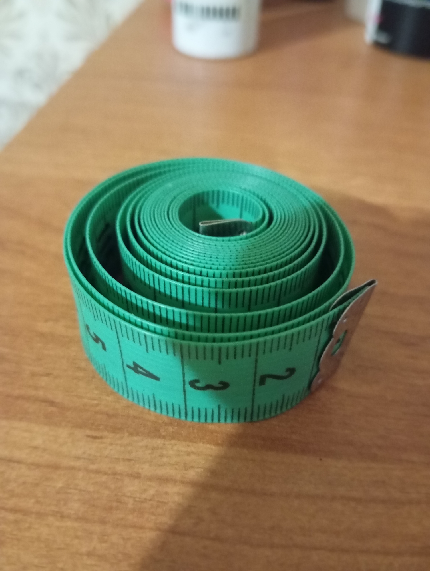 The image displays a coiled green measuring tape on a wooden surface. The measuring tape has black markings and numbers, with visible numbers 2, 3, and 4. The background is blurred, showing a light-colored wall and some out-of-focus objects, one of which appears to be a white cylindrical container with black text. There is no indication of the location being Orenburg, Russia, or any city for that matter. No people or actions are depicted in the image.