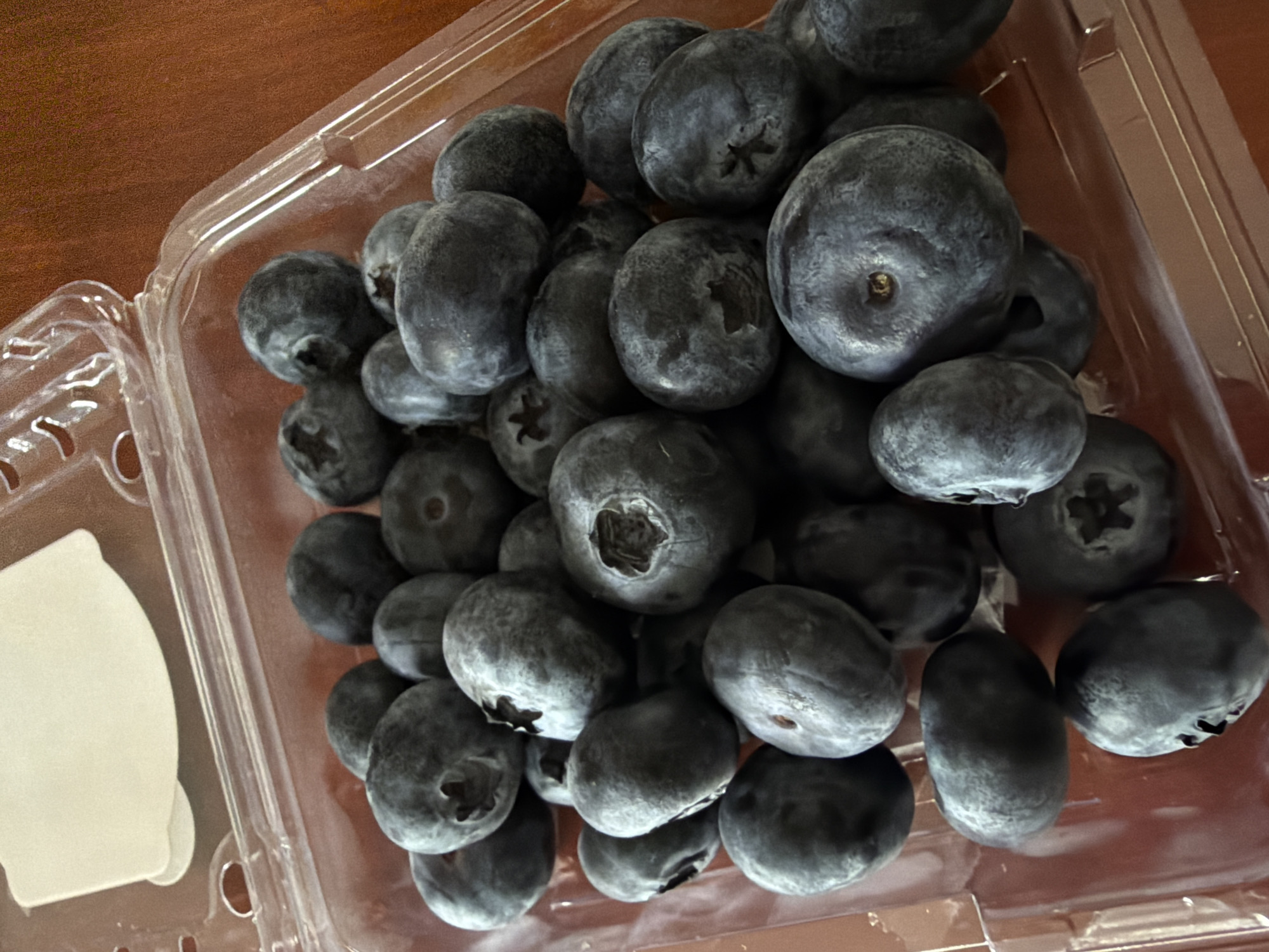 This is a top-down, close-up shot of a clear plastic container filled with fresh blueberries. The blueberries are a deep blue color with a slightly dusty, matte finish, indicating they are ripe. They are densely packed within the container, with some spilling over the edges. The container itself is clear plastic with a textured bottom and a hinged lid that is partially open, revealing the contents. The background is a warm, brown wooden surface, likely a tabletop. The lighting is bright, suggesting it could be daytime, but there are no shadows that would indicate a specific time of day. The scene is simple and focuses entirely on the blueberries, conveying a sense of freshness and abundance. There is no visible text in the image. The overall impression is one of healthy, natural food presented in a straightforward manner. The location context, Chang Phueak, Thailand, is not visually discernible from the image itself.