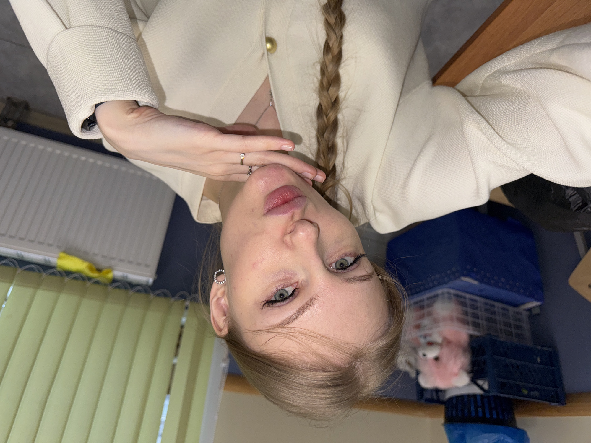 A light-skinned young woman with a blonde braid is posing indoors. She is wearing a white jacket. Her left hand is near her chin, displaying a ring on her finger. The background includes vertical blinds, a radiator, a blue storage container with a pink stuffed animal, and what appears to be a wooden chair. The location appears to be in a room, inside a building in Perm, Russia.