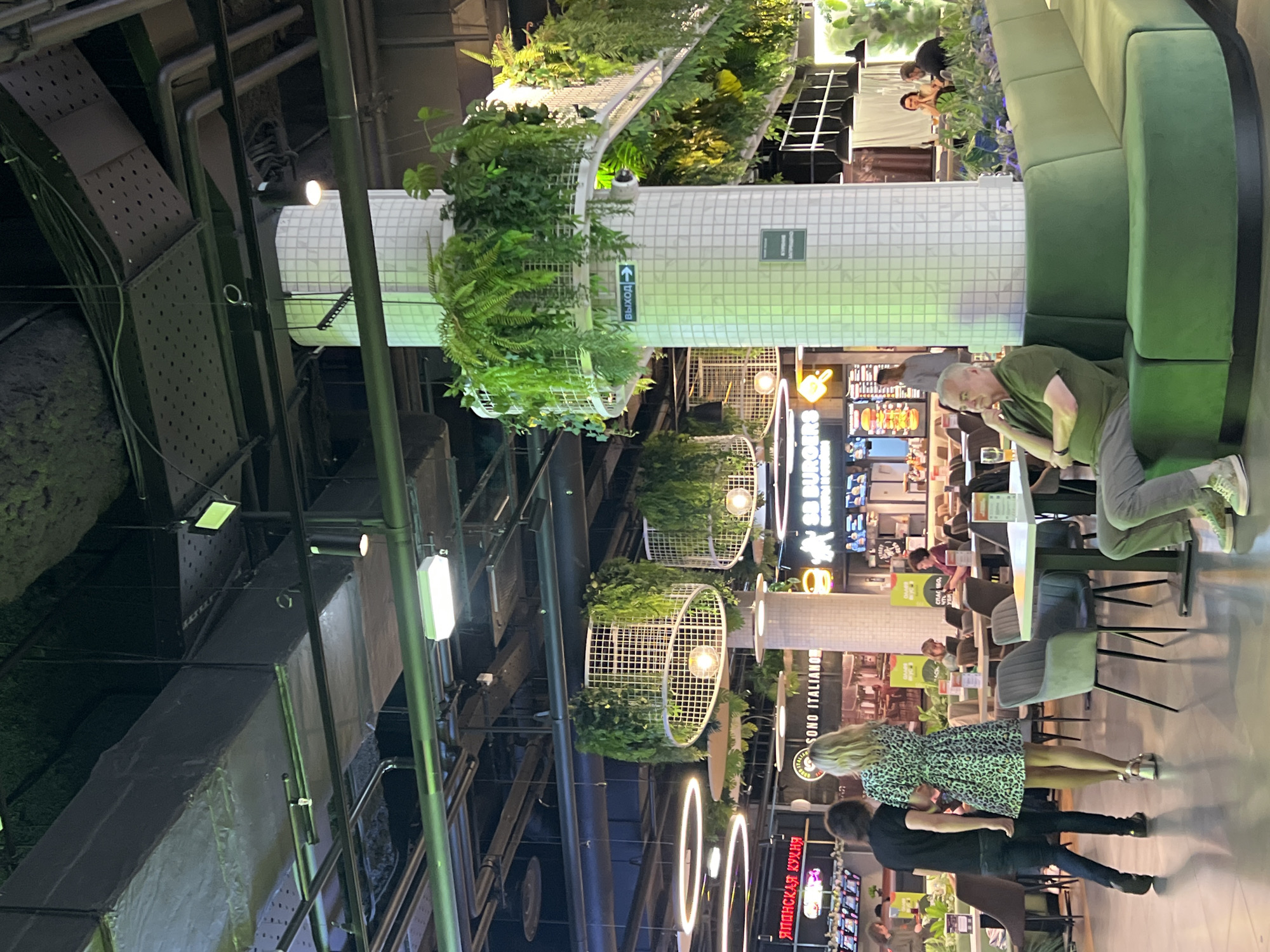 This image depicts an interior scene, possibly a food court or a restaurant area within a large building, given the presence of multiple dining establishments and seating arrangements. The environment appears to be a modern indoor space with a high ceiling and industrial-style exposed infrastructure, including pipes and ventilation systems. Decorative elements include hanging plants and circular lighting fixtures, creating a blend of natural and contemporary aesthetics.
Several people are visible in the scene, engaged in various activities. In the foreground, a man in a green t-shirt and grey pants is seated, looking down. Behind him, a couple is standing, with the woman wearing a patterned dress and the man in a dark shirt. Further in the background, other individuals are seen dining or walking. The lighting suggests it is likely evening or indoors with artificial illumination.
Visible text includes signs for "BURGER" and "ЯПОНСКАЯ КУХНЯ" (Japanese Cuisine) in Russian, and "SONO ITALIANO" (possibly meaning "It's Italian" or "Sono Italiano Restaurant") in English and Italian. There is also a sign with an arrow and the Russian word "ВЫХОД," meaning "Exit." These cues indicate a diverse food court or restaurant setting. The overall mood appears casual and social, typical of a public dining area. The location context of Ladozhskoe Ozero, Russia, further supports the interpretation of a Russian establishment.
