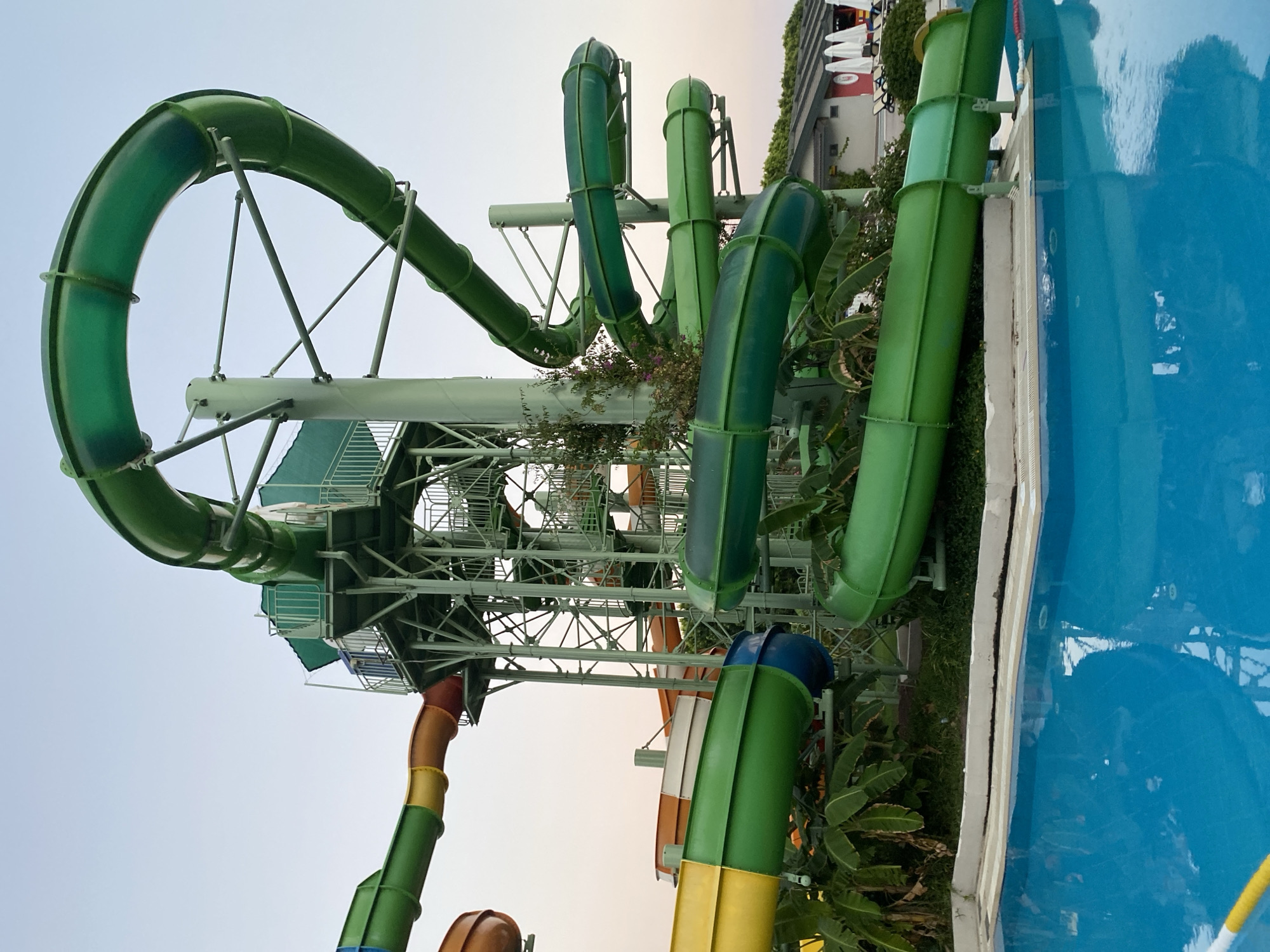 An exterior shot captures a vibrant water park attraction in Aksu, Turkey, under a clear, pale sky. The central focus is a multi-level, green water slide structure with numerous winding tubes and a tall, metallic support tower. One section of the slide forms a large green loop, while other parts twist and spiral downwards. Several slide segments are visible in various colors, including green, yellow, orange, and red, suggesting a complex network of rides. Lush green banana plants and other foliage surround the base of the slides, bordering a bright blue swimming pool. In the background, a low-rise building with a light-colored roof and several white umbrellas is visible, hinting at a resort or leisure facility. The scene is devoid of people, emphasizing the architectural elements of the water park.