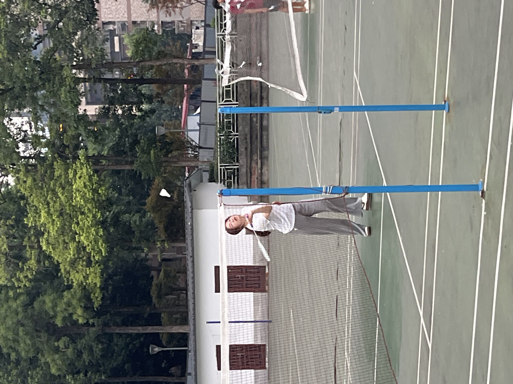 This image depicts a badminton court in Yuzhong District, China, with two individuals actively engaged in a game. A woman in the foreground, wearing a white striped shirt and gray pants, is in mid-swing, her racket raised to strike a shuttlecock that is visible in the air. She appears to be focused on the game, with her head tilted back and eyes looking upwards. To her right, partially obscured by the net, another person in a red shirt and dark shorts stands ready to play. The court is surfaced with a green, textured material marked with white lines, indicating boundaries for the game. A white net with blue poles divides the court. In the background, a white building with two dark doors is visible, along with a row of trees and some urban buildings in the distance. The overall scene suggests an outdoor recreational activity taking place on a cloudy day.
