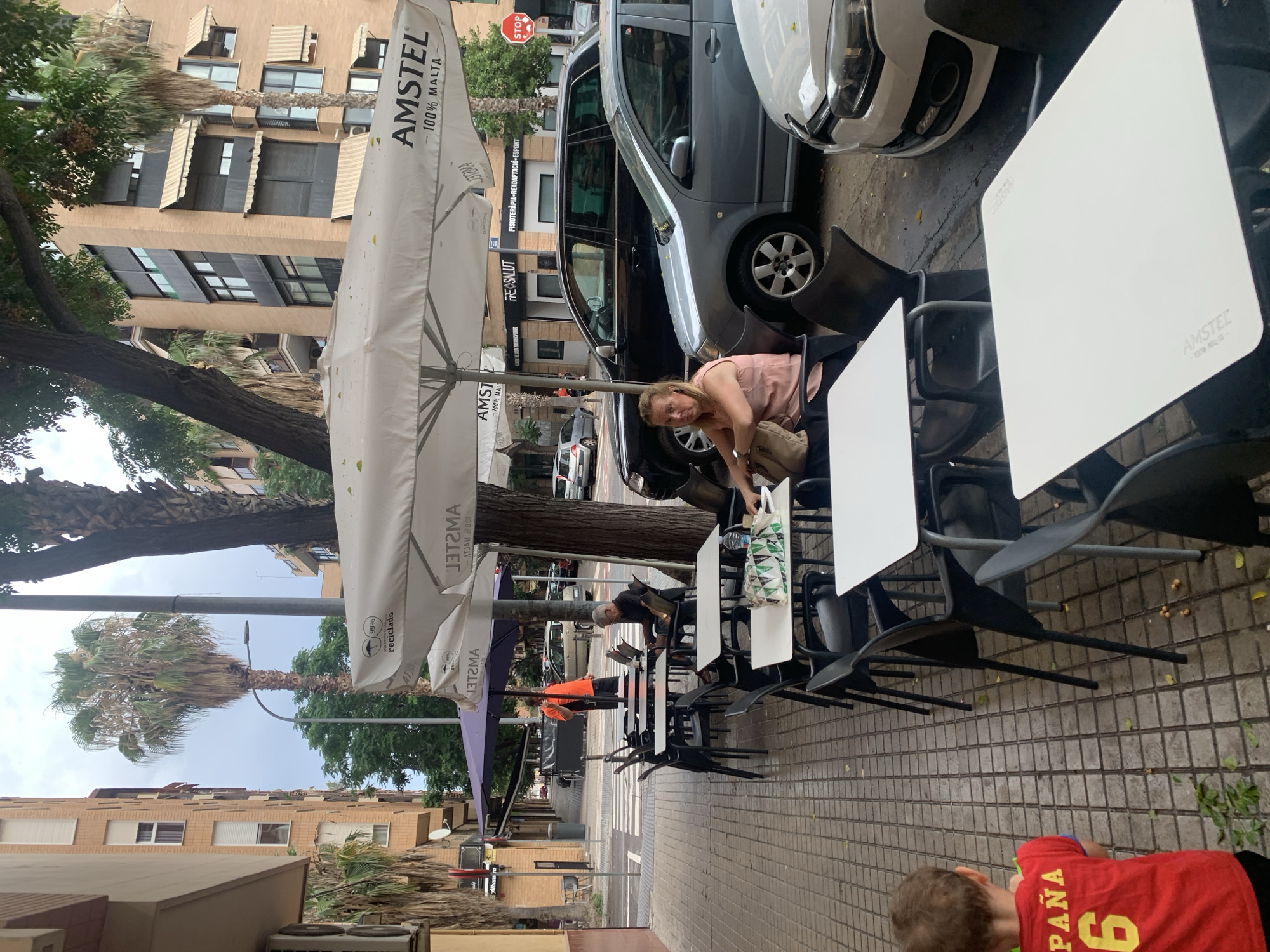 The image depicts an outdoor seating area of a cafe or restaurant located on a street in Valencia, Spain. The scene is set on a tiled sidewalk with several tables and chairs. In the foreground, a young boy in a red shirt with "España 6" on the back is walking away from the camera. Behind him, a woman with blonde hair is sitting at one of the tables, looking towards the camera. She is wearing a pink top and carrying a beige handbag. Further down the sidewalk, a man in an orange shirt is standing near a purple umbrella, and another man in a black shirt is seated at a table. Several cars are parked along the street, including a black SUV and a grey sedan. The cafe's umbrellas are branded with "AMSTEL 100% MALTA." The buildings in the background are typical of urban architecture in Spain, with balconies and awnings. Palm trees are visible, suggesting a Mediterranean climate. The overall impression is a casual outdoor dining environment on a city street.