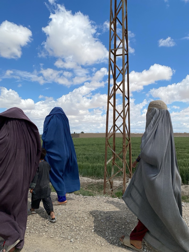 The image shows three women and a child walking along a gravel path near what appears to be a field in Mandisar, Afghanistan. The women are dressed in full-body coverings, one in purple, one in blue, and one in gray. A child in a dark-colored outfit walks between the purple and blue figures. An old metal tower stands to the right of the blue figure, close to the gray figure. The scene takes place under a blue sky with fluffy white clouds. A green field stretches into the horizon, and a building is visible on the far-right.