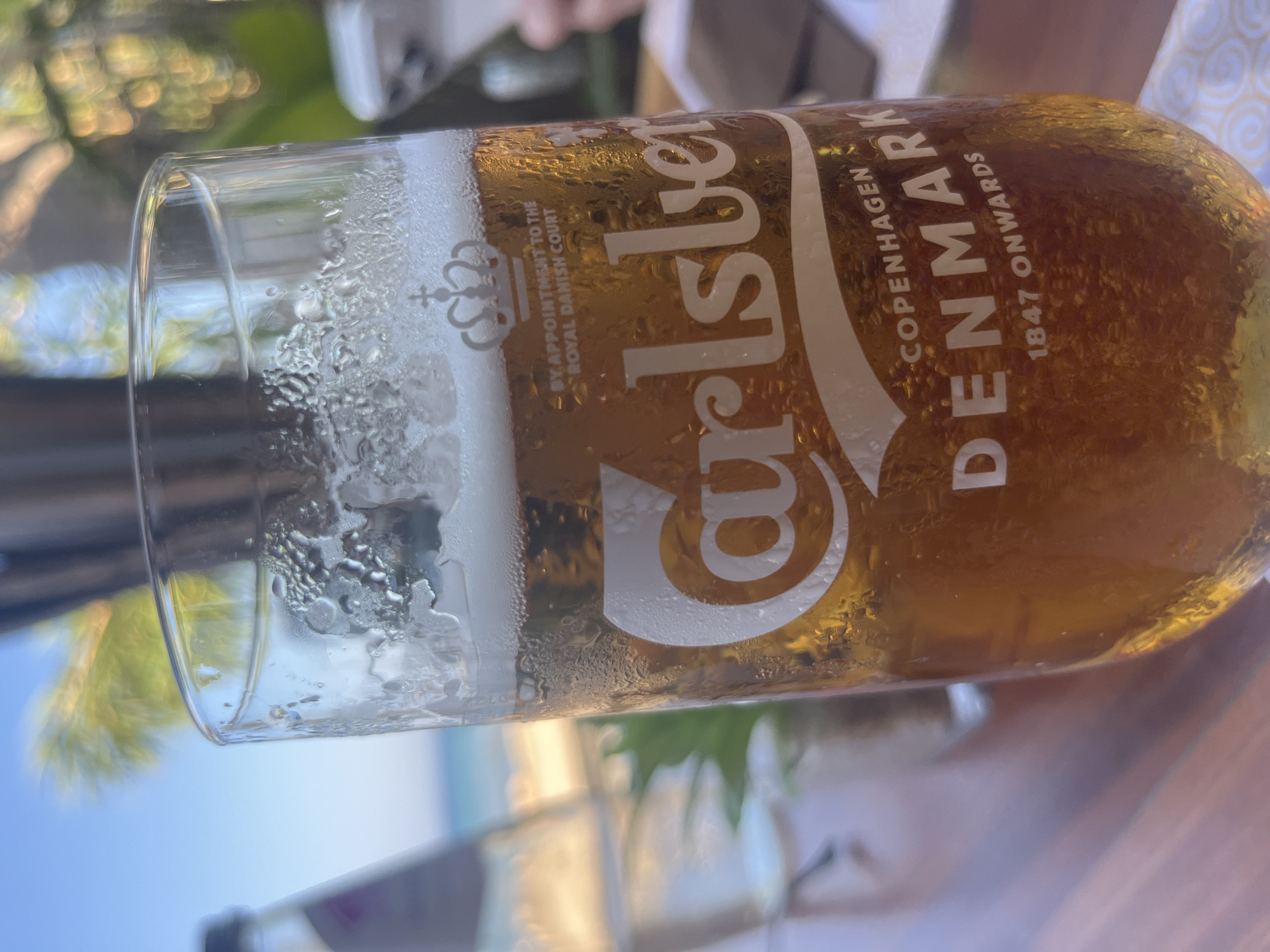 The image is a close-up shot of a glass of Carlsberg beer, condensation visible on the exterior, placed on a wooden table outdoors. In the background, blurred elements suggest a coastal setting with a body of water and possibly palm trees, consistent with a location like Protaras, Cyprus. The beer glass displays the Carlsberg logo, the royal Danish crown, "BY APPOINTMENT TO THE ROYAL DANISH COURT", "COPENHAGEN DENMARK", and "1847 ONWARDS". A bottle of water is visible to the left, and a hand holding a mobile phone is partially seen on the right. The lighting indicates it is daytime, possibly late afternoon due to the warm tones.