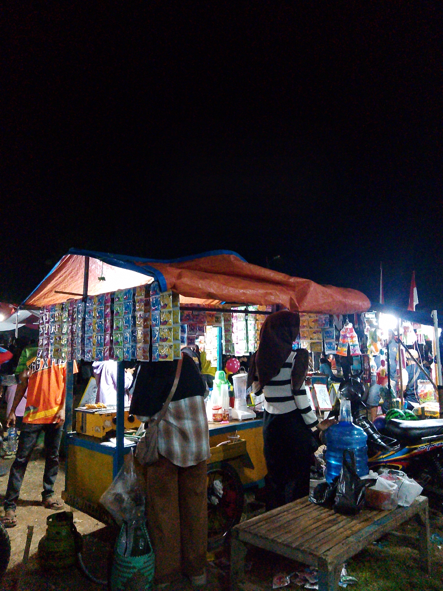 A nighttime outdoor scene in Wanareja, Indonesia, features a street food stall with various items for sale. A vendor, wearing a brown hijab and a black and white striped top, stands behind the stall. To the left, a person in an orange shirt and blue jeans is visible, holding a plastic bottle. The stall itself is covered by an orange tarp and displays numerous packets of colorful goods hanging on the front. A yellow cart serves as the base for the stall, with a grill-like apparatus on top where food is being prepared. In the foreground, a wooden bench is placed in front of the stall, with a large blue water jug and several plastic bags on it. The ground is covered in dirt and grass, and there are other people and stalls visible in the background. Two Indonesian flags are also seen in the distance. The overall atmosphere appears to be that of a lively night market or fair.