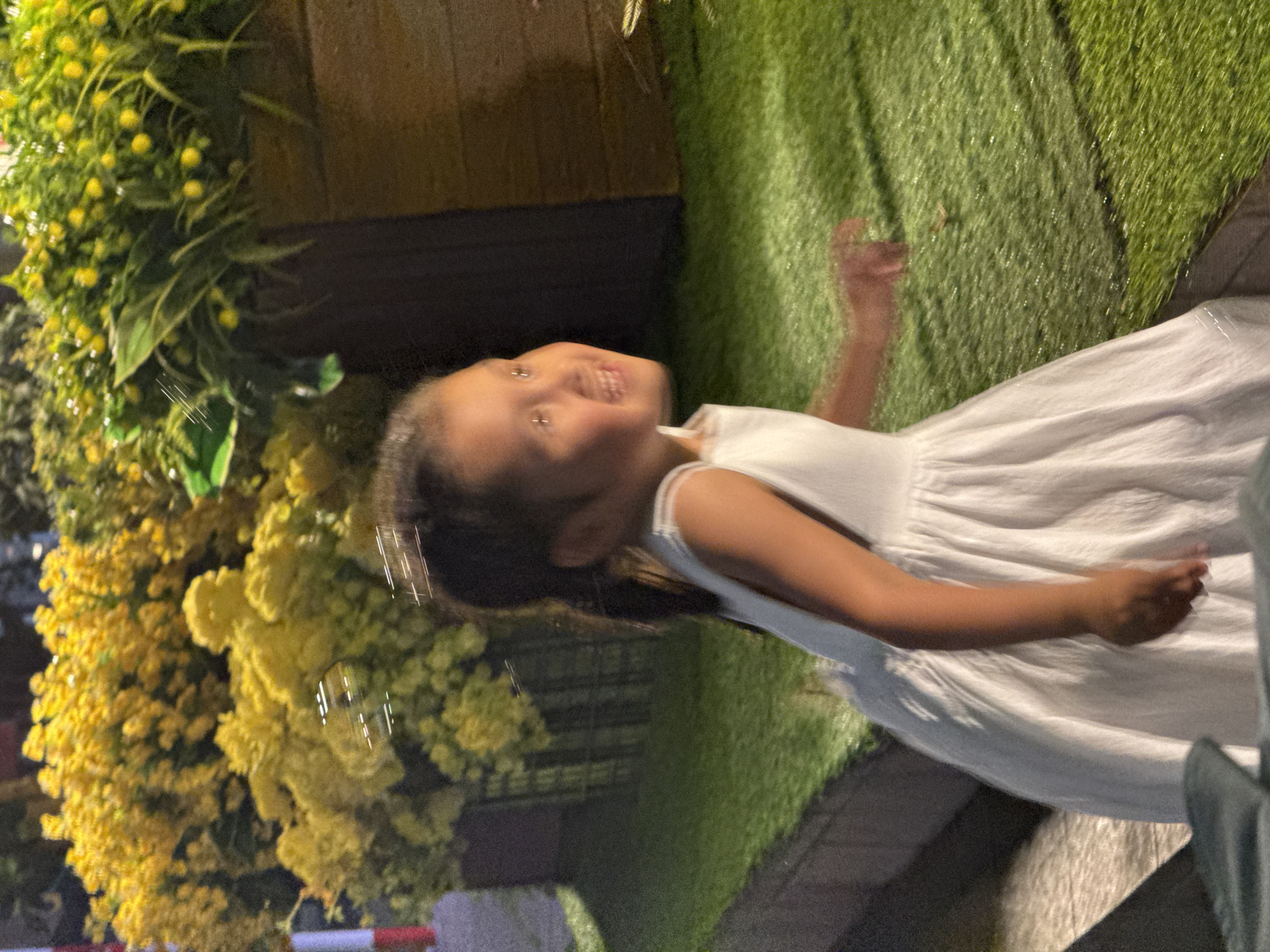 This image captures a joyful young girl in an outdoor or semi-outdoor decorated setting, likely in Nha Trang Ward, Vietnam. The girl, appearing to be of preschool or early elementary age, wears a white dress and has dark hair. She is depicted mid-action, smiling broadly and looking upwards and slightly to her left, with her hands raised in a playful gesture.
The background on the left features dense, vibrant green foliage interspersed with clusters of bright yellow artificial flowers, possibly mimosa or similar decorative plants. Darker wooden structures are visible behind the foliage. To the girl's right, there is green artificial turf or grass. Several small, reflective, spherical objects, resembling soap bubbles, are visible floating in the air near her face and among the foliage, suggesting she is engaging in play with bubbles.
The lighting indicates it is evening or night, with artificial illumination highlighting the scene. The girl's wide smile and animated posture convey feelings of happiness, excitement, and childlike play. No visible text is present in the image.