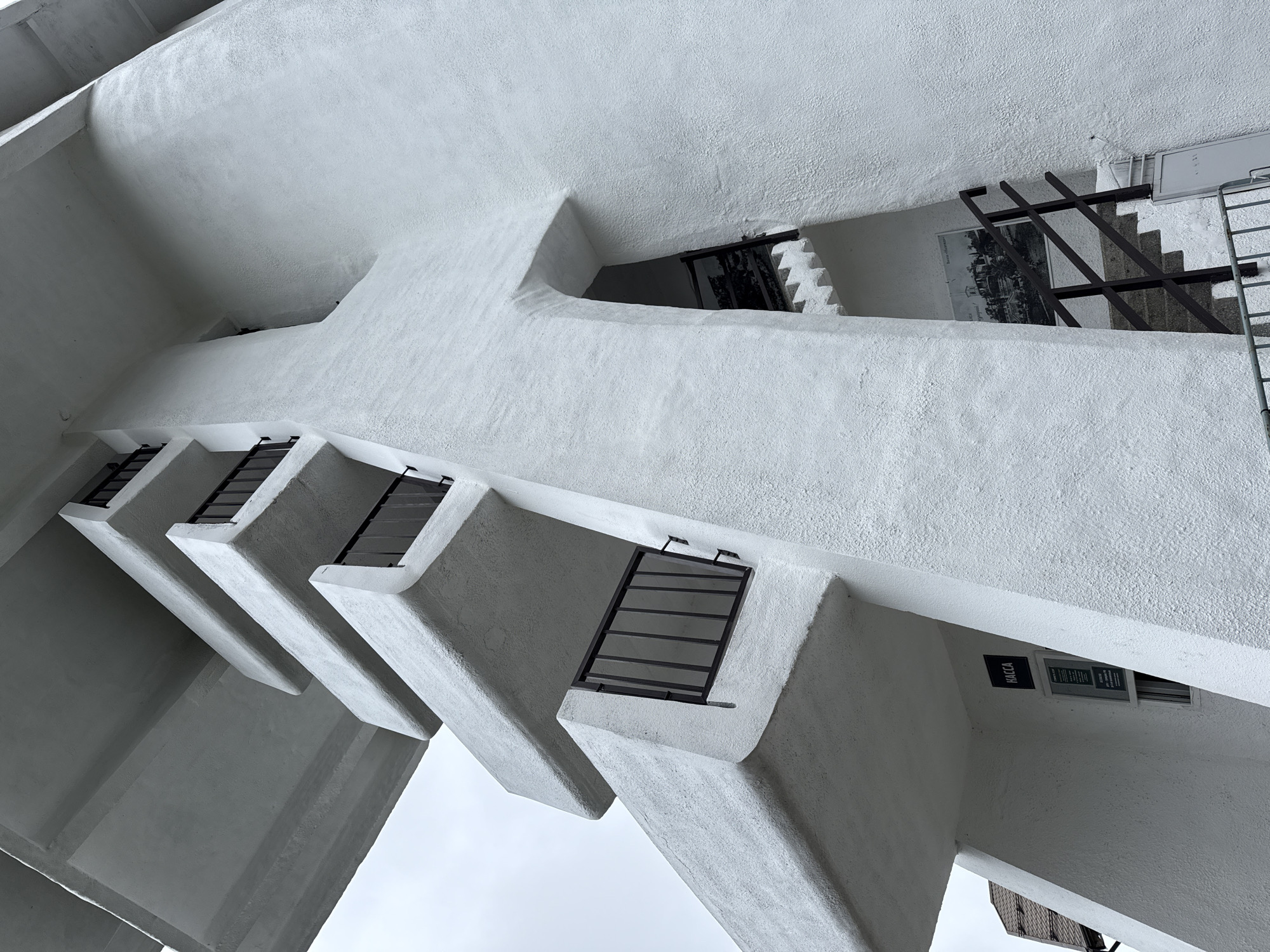 This low-angle photograph depicts the Mount Akhun Observation Tower, located near Sochi, Russia. The structure is an example of Stalinist-era modernist architecture, characterized by its stark white, textured concrete or stucco exterior. Multiple levels of sharply cantilevered, rectangular viewing platforms or balconies project outward, each secured with dark, vertically-barred metal railings. A prominent, angular central vertical element forms the tower's core, creating a distinctive 'cross' or 'T' shape in profile against an overcast, gray sky. At the lower left of the structure, a dark square sign displaying the Cyrillic word "KACCA" (Cashier/Ticket Office) is mounted above a small window with a framed notice. An arched entryway is visible into the tower's base, revealing a set of dark-colored stairs with metal handrails ascending into the interior. A black and white historical photograph is visible on the inner wall of the stairwell. No individuals are present in the scene.