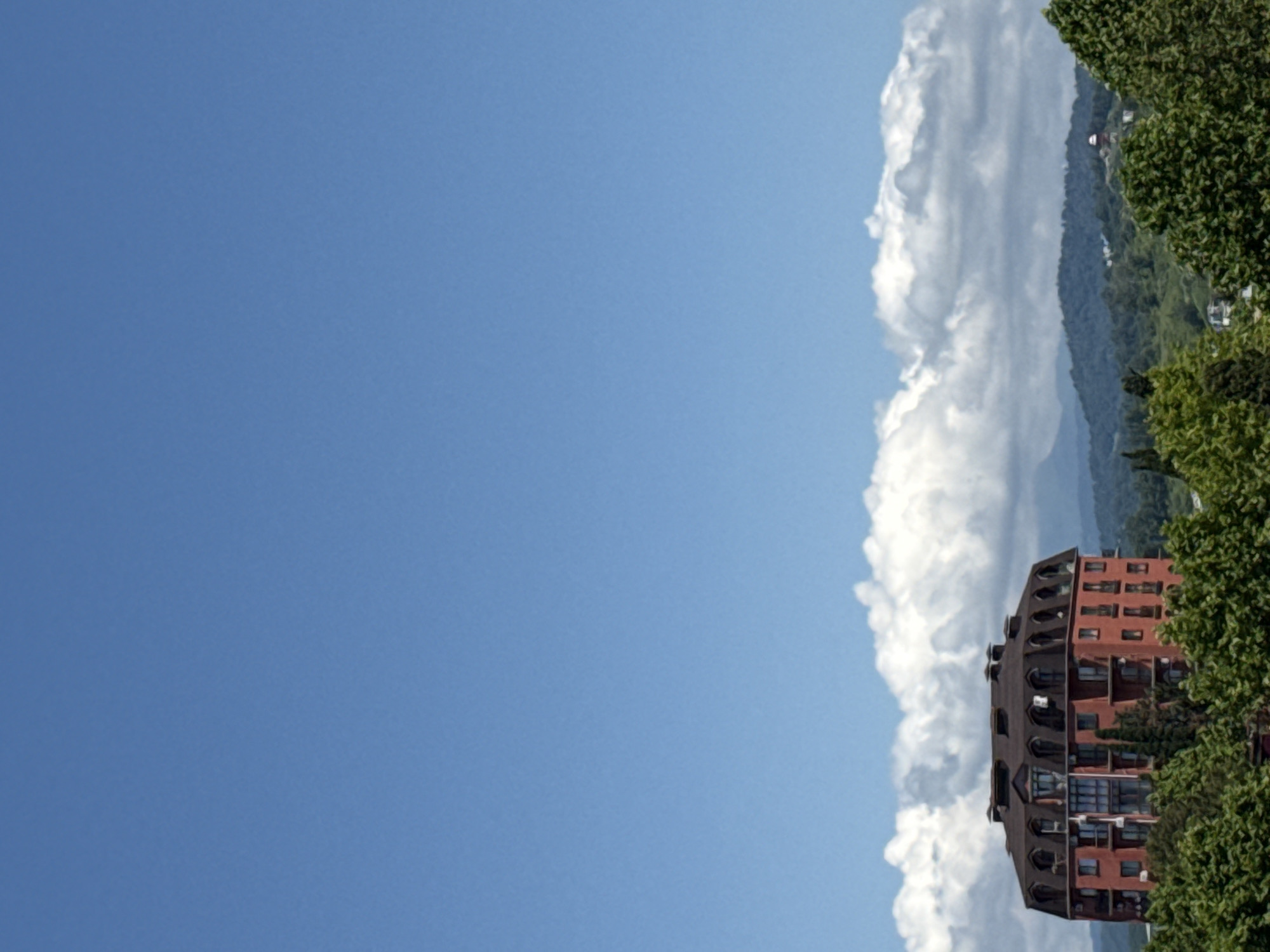 This image depicts a contemporary, red-brick building with a distinctive mansard roof featuring numerous dormer windows, situated amidst lush green trees. The building appears to be located in the city of Sochi, Russia, given its architectural style and the surrounding hilly, verdant landscape, which is characteristic of the region. In the background, rolling hills are visible, topped with a line of fluffy white clouds against a clear blue sky. The scene suggests a bright, sunny day. No people or specific actions are discernible in the photograph. The focus is on the building and its natural surroundings.