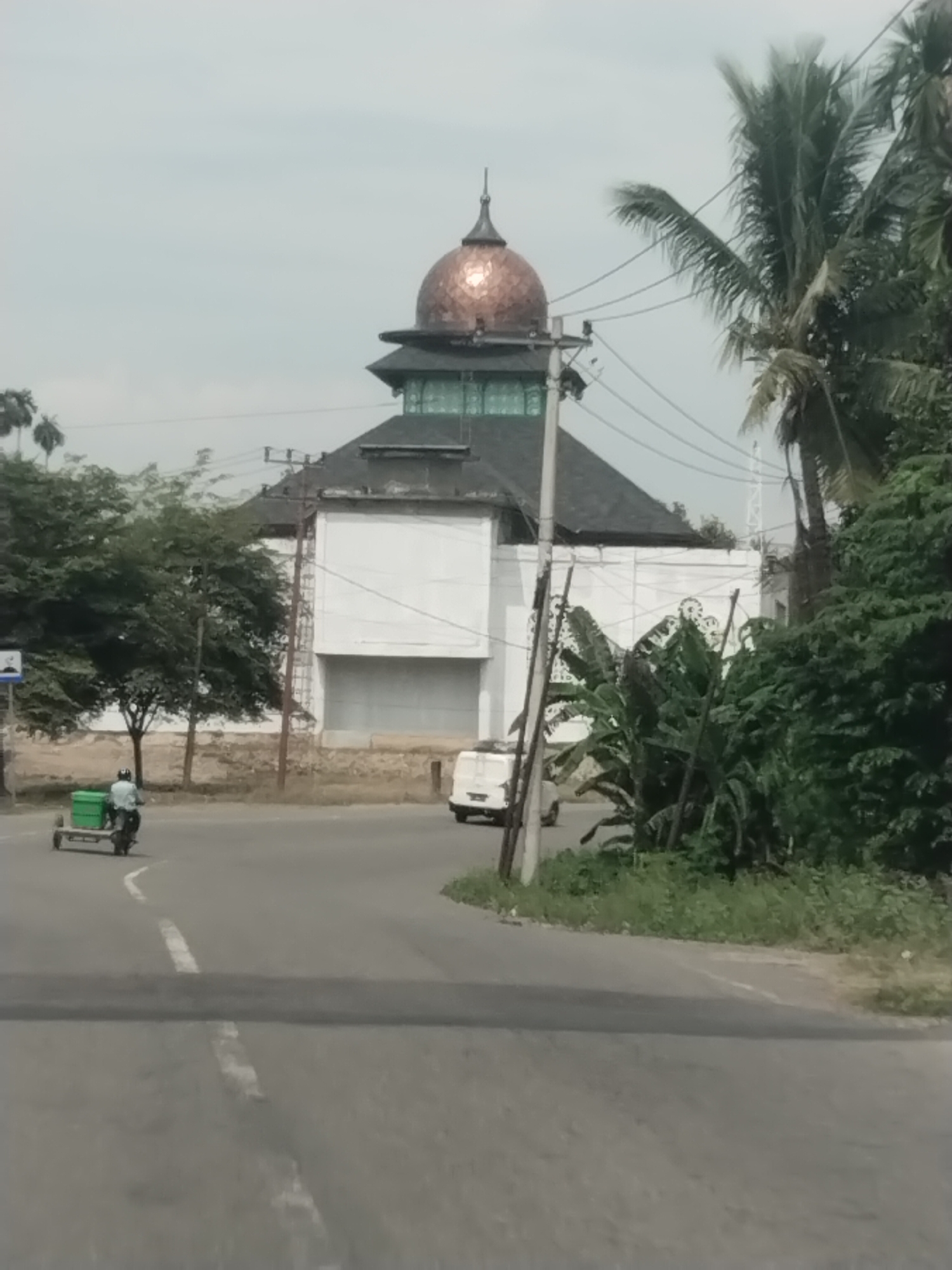 The image shows a street scene in Manyang Cut, Indonesia, with a mosque in the background. A motorcycle with a sidecar carrying two green containers is traveling on the paved road, which has white lane markings. To the right of the road, there is lush green vegetation and palm trees. The mosque features a prominent copper-colored dome and dark roof. A white van is parked on the side of the road near the mosque. The sky is overcast, indicating a cloudy day. The overall environment appears to be a semi-urban or rural setting.
