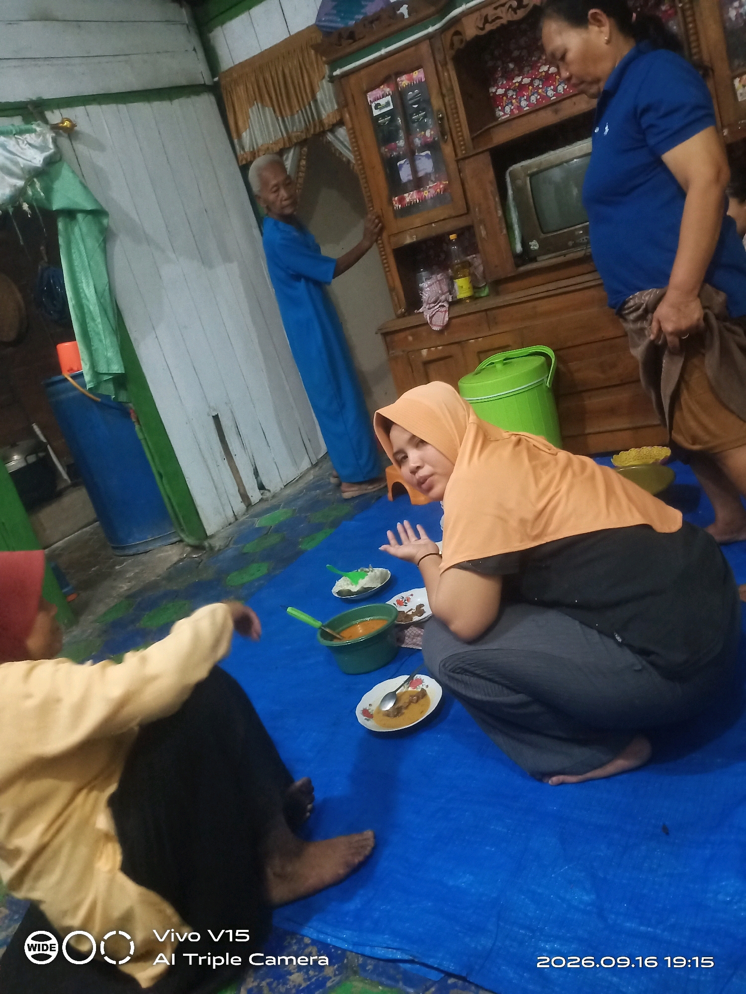 In Plosorejo, Indonesia, a domestic scene unfolds within a modestly furnished room, likely a home. The setting is characterized by wooden walls and a rustic wooden cabinet. A blue tarp covers the floor, indicating a casual gathering or meal.
Four individuals are present. An elderly man, wearing a blue shirt, stands near the cabinet, his attention directed away from the camera. Beside him, a woman in a blue polo shirt and a patterned sarong is also standing, her gaze lowered. In the foreground, a woman wearing an orange hijab and grey pants is seated on the tarp, looking towards the camera. To her left, another person, partially visible and wearing a yellow top and red headscarf, is also seated.
The table is set with plates of food, suggesting a meal is in progress. A large green plastic container is also visible. The lighting appears to be artificial, contributing to a dimly lit atmosphere. The overall impression is that of a family or community gathering, possibly for a meal.