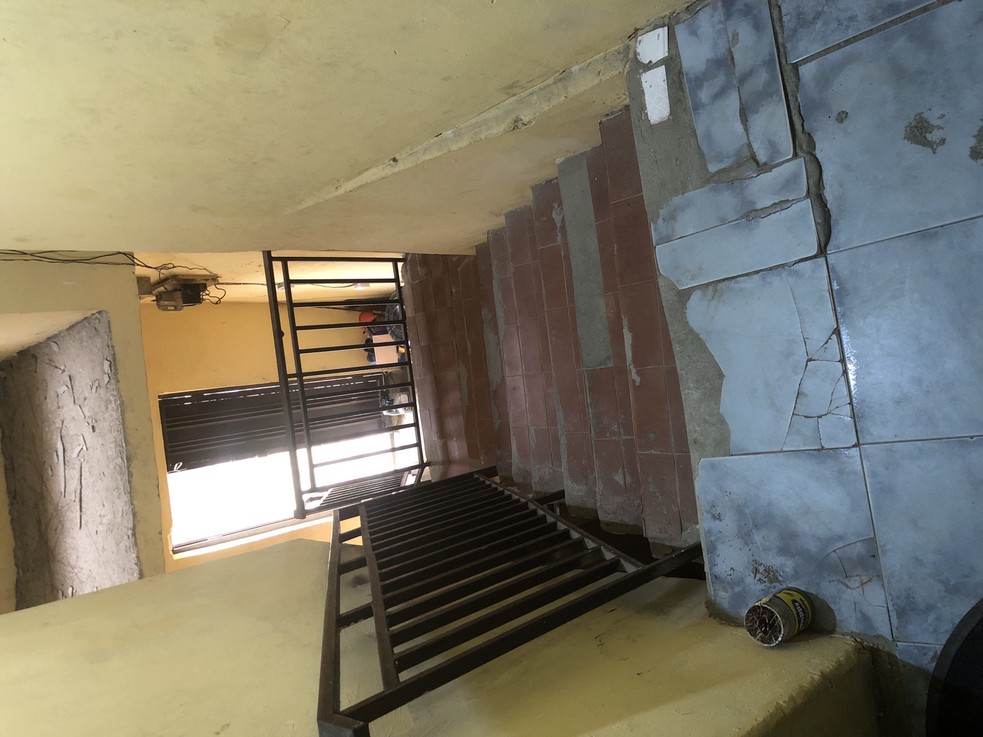 This is a photograph taken in Ado, Nigeria, likely within a residential building or a public staircase. The image shows a descent down a staircase, with yellow-painted walls on either side. The staircase itself is made of concrete steps, with some steps tiled in a reddish-brown color and others covered in light blue tiles that appear cracked and unevenly laid. A dark metal railing runs along the left side of the stairs, with vertical bars and a horizontal top rail.
At the top of the staircase, a doorway is visible, leading to an area with bright natural light. In front of the doorway is a black metal gate or barrier. Some items, possibly boxes and other miscellaneous objects, are visible behind the gate. An electrical meter with wires is mounted on the yellow wall to the right of the doorway.
On the left side of the staircase, near the bottom of the frame, a small cylindrical container filled with what appears to be discarded matches or similar items rests against the yellow wall on a tiled surface. The overall impression is of an aged and somewhat neglected urban interior space.