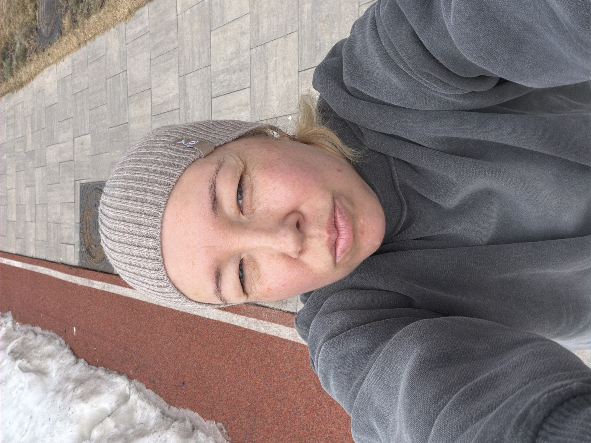 A woman wearing a grey beanie and a grey sweatshirt takes a selfie outdoors. The ground is a combination of concrete paving stones, a red track with a white dividing line, and a patch of snow. In the background, there's a manhole cover. The location is Pervomaysky, Russia.