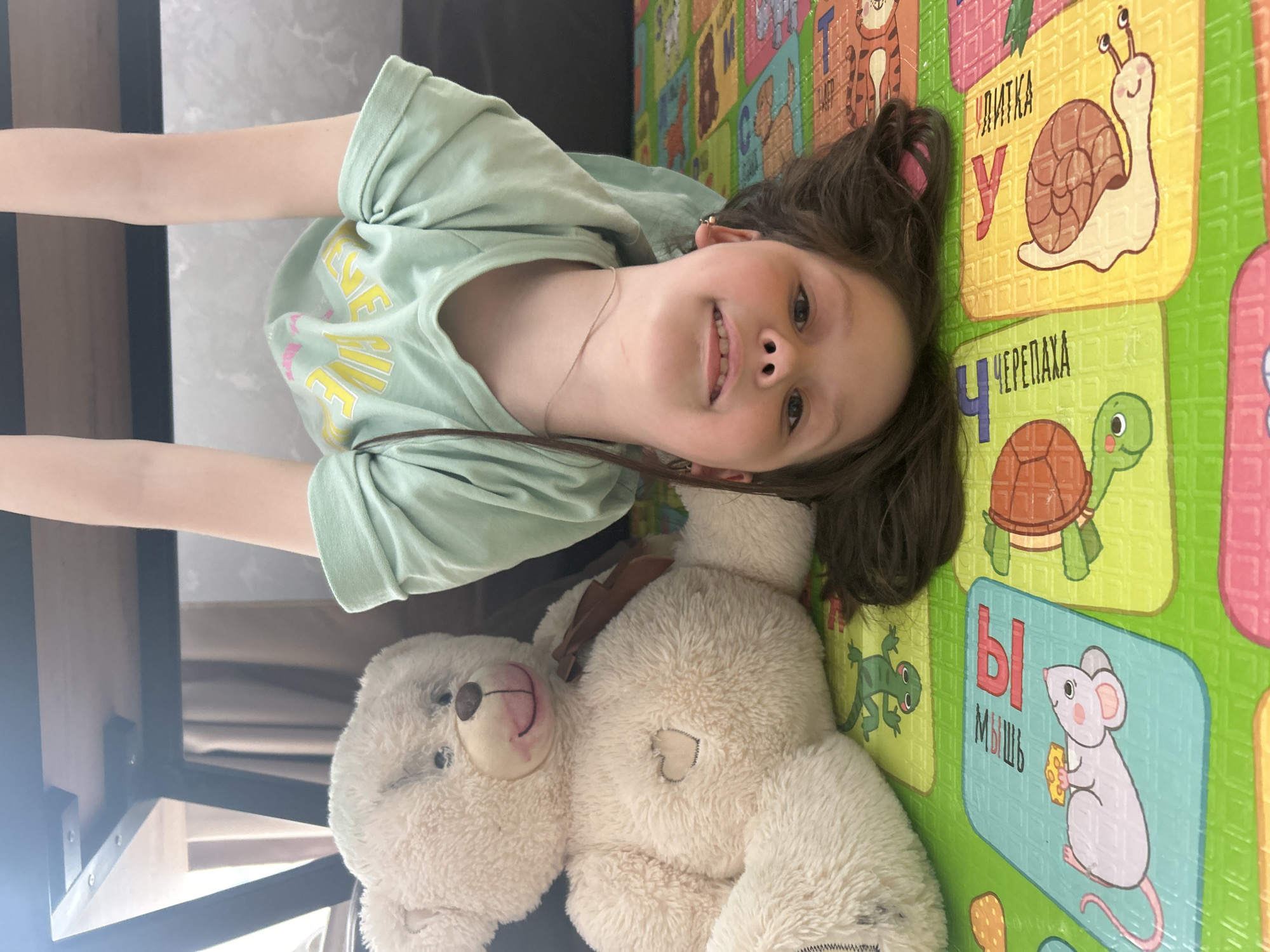 A young girl lies on a colorful play mat next to a white teddy bear. She wears a light green t-shirt and has brown hair pulled back with a pink hair tie. She is smiling, her arms stretched out to either side. The play mat features Russian alphabet letters and corresponding animal illustrations (snail, turtle, mouse). The background includes glimpses of furniture legs and neutral-toned curtains. It is not possible to determine the specific location in Chaikovsky, Russia from the image.
