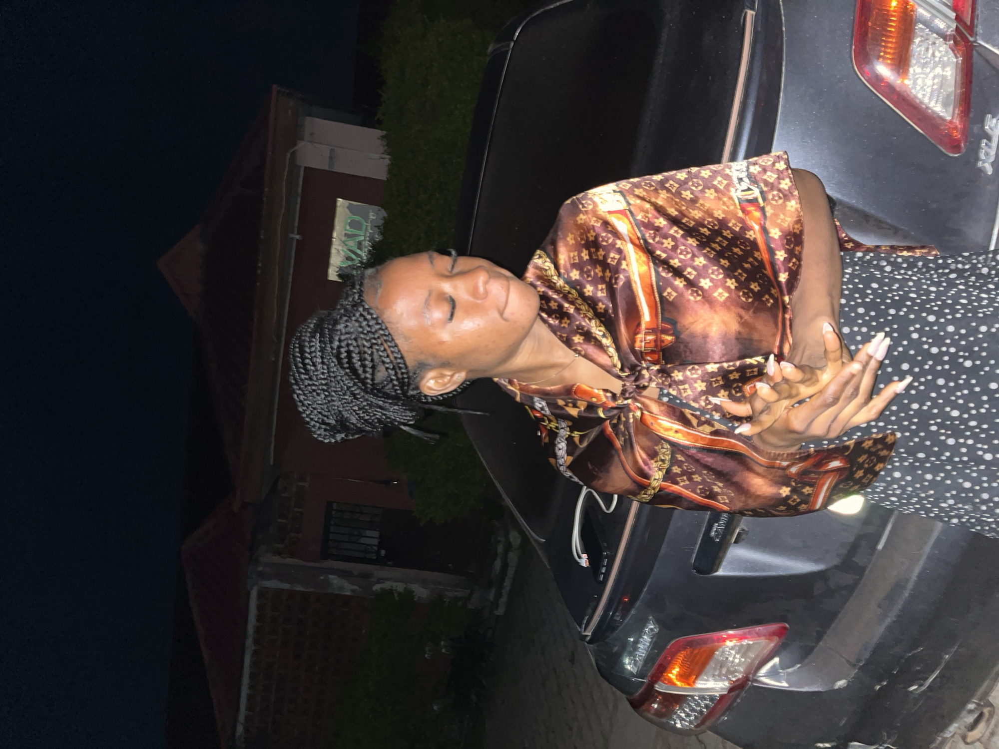 A young Black woman with braided hair is standing next to a dark-colored car at night, with a building in the background. She is wearing a patterned silk top and a polka-dot skirt. The building appears to be a house with a red roof. The scene is dimly lit, suggesting it is nighttime. There is a phone and a white cable on the trunk of the car. The image appears to have been taken in an urban or suburban area.