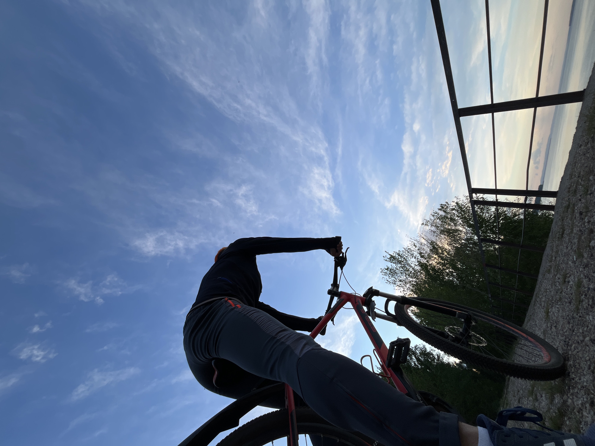 A low-angle, outdoor shot shows a person on a red mountain bike against a backdrop of a blue sky with cirrus clouds and a view of water.
The person is wearing dark clothing and light gray pants and is seated on the bike, holding the handlebars. Only parts of their feet and shoes are visible. The red bike is angled towards the top right of the frame.
To the right, a metal railing or barrier is partially visible, with a view of water beyond it. Beneath the barrier are trees and some concrete.
Given the prompt, it can be inferred that the location is near water with trees, with a possible overlook or barrier in the city of Chaikovsky, Russia.