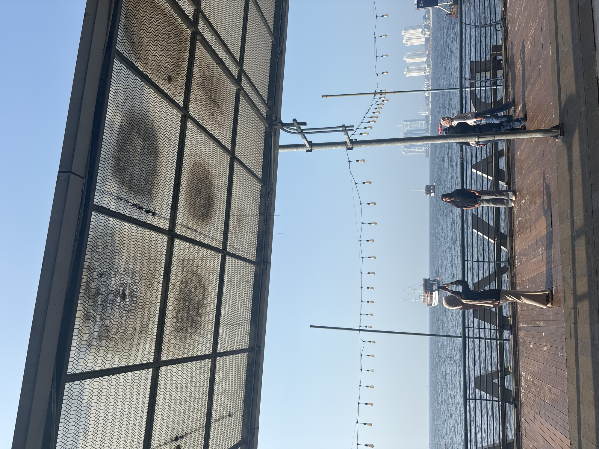 This image depicts a scene at Ancol Beach City, North Jakarta, Indonesia, on a clear day. In the foreground, a wooden boardwalk extends into the frame, leading to a metal railing that separates the walkway from the water. A decorative string of lights hangs above the boardwalk, and a metal canopy with a mesh covering provides shade.
Several individuals are present on the boardwalk. Closest to the camera, a person wearing a hijab and a beige outfit is standing and taking a photo with a smartphone, facing the water and a distant boat. To their right, another person in a dark top and jeans stands facing the camera. Further to the right, two more individuals are visible, one wearing a hijab and the other in a dark top and jeans, both facing the water.
The sea is calm and stretches to the horizon, where a large boat with sails is visible in the middle ground. In the background, a city skyline with tall buildings and cranes suggests the urban environment of North Jakarta. The overall atmosphere is relaxed and casual, with people enjoying the waterfront.