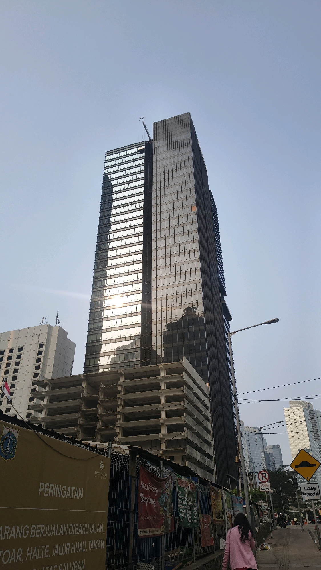 A low-angle shot captures a modern skyscraper under construction in the city of South Jakarta, Indonesia. The building, clad in reflective glass, displays a prominent construction crane at its apex. Adjacent to it, an unfinished concrete structure with visible floors and supports indicates ongoing development. A tall, white building with rectangular windows stands to the left of the main skyscraper. The foreground features a construction fence adorned with various banners, one with yellow text reading "PERINGATAN" and another with Indonesian text. A pedestrian walkway lines the right side of the image, where a young woman in a pink jacket walks away from the camera. Streetlights and traffic signs are visible along the walkway, including a speed bump warning sign and a prohibition sign. The sky is clear and pale blue, suggesting a daytime scene.