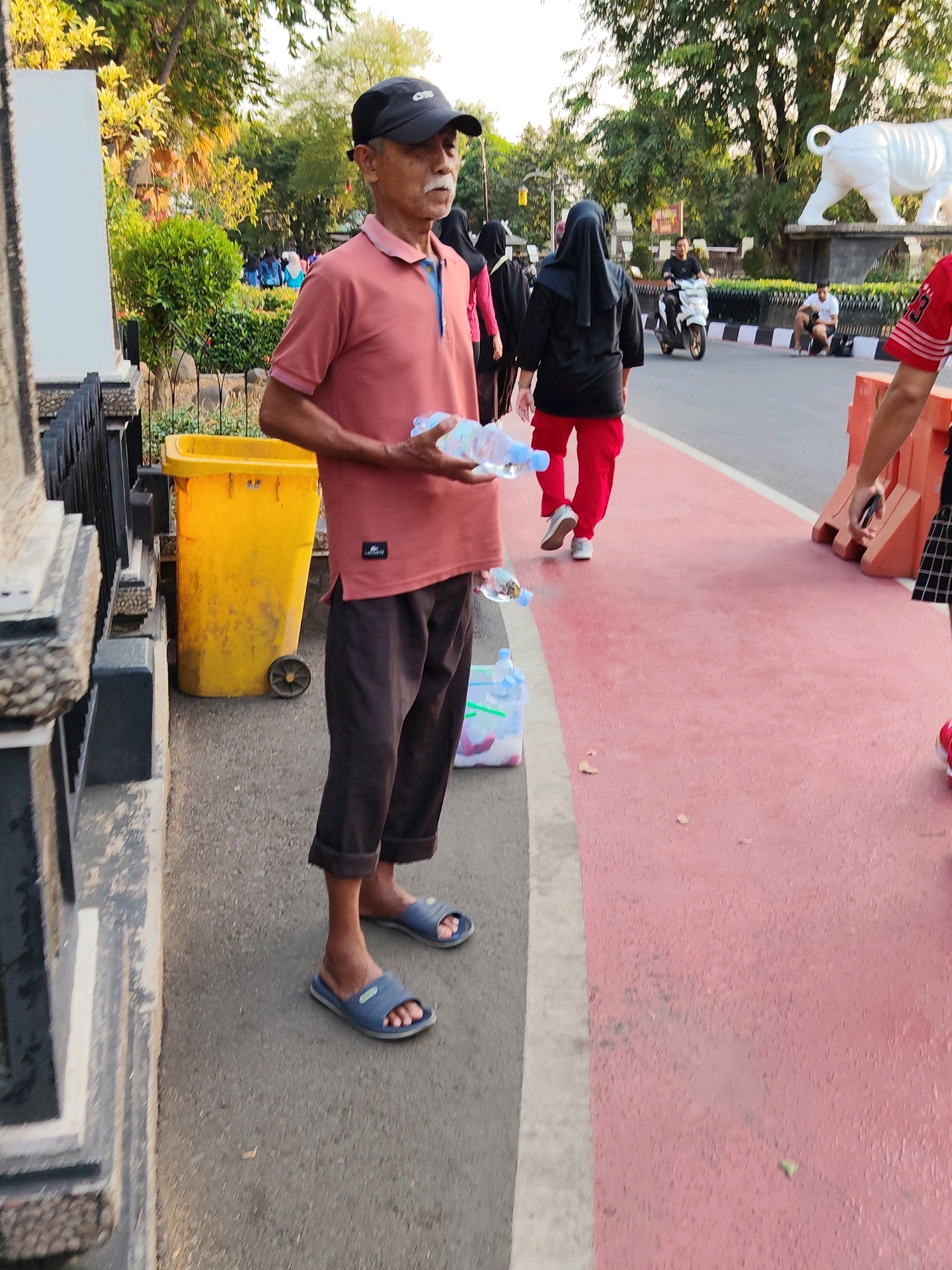 In Purwakarta, Indonesia, an elderly man wearing a pink polo shirt and dark pants, with a black baseball cap, stands on a sidewalk. He holds two plastic water bottles. In the background, a bright yellow trash bin with wheels is visible. Further back, several people are walking, one wearing a black hijab and red pants. A motorcycle passes by, and a white sculpture of a rhinoceros or similar animal is present in the distance. The ground is paved, with a red-colored lane or path next to the sidewalk. The scene appears to be an outdoor public space, possibly a park or recreational area, with trees and foliage contributing to the environment.