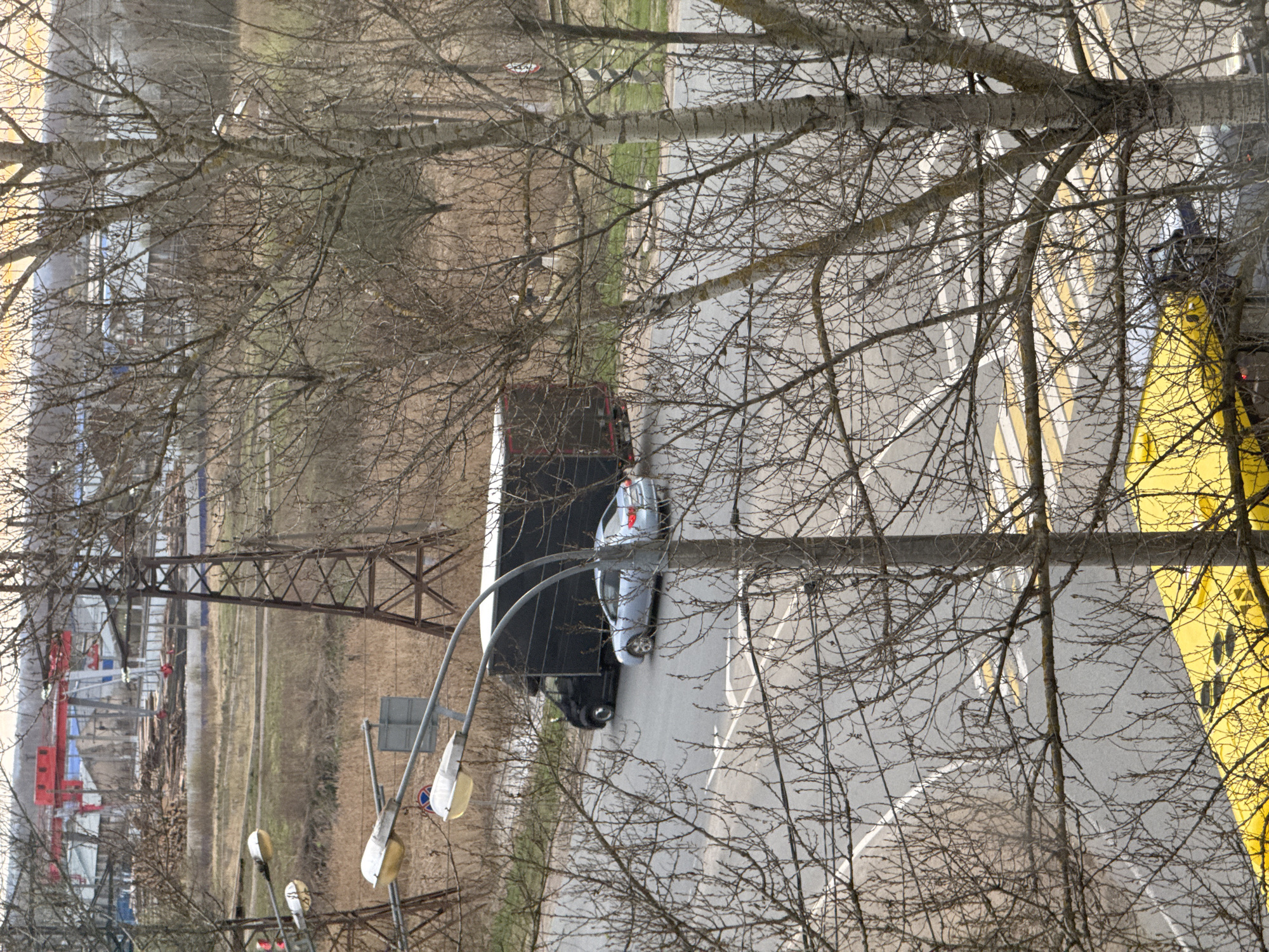 The image presents an elevated view overlooking a road, bare trees, and an industrial or construction area, likely during late autumn or early spring due to the leafless trees. The scene is lit by diffused daylight, suggesting an overcast sky or hazy conditions.
In the foreground, a dense network of bare tree branches frames the view. Below these branches, a paved road stretches across the middle ground. On the road, a dark-colored truck, possibly a large delivery vehicle or a semi-truck cab, is partially visible, followed by a light-colored sedan (silver or light blue). Road markings, including solid white lines and diagonal yellow lines, are discernible. To the right of the road, a prominent, bright yellow structure or banner with dark markings, possibly a biohazard symbol, takes up a significant portion of the frame. Streetlights with multiple lamps are visible on the left side of the road.
Beyond the road and trees, a wide, open field with dry grass extends into the distance. In the far left-middle ground, a large, dark brown metal truss structure, possibly part of a bridge or industrial gantry, spans across the landscape. Further left, in the background, red and blue industrial equipment, resembling cranes or parts of a factory, are visible, alongside piles of what appears to be construction material or debris.
No people are explicitly visible, but the vehicles on the road imply human presence and activity. The overall setting combines elements of urban infrastructure, natural landscape, and industrial development, consistent with an area like Novonikolskoye, Russia. No clear, readable text is present, aside from the ambiguous markings on the yellow structure and a partially visible circular traffic sign with a blue background and red border.