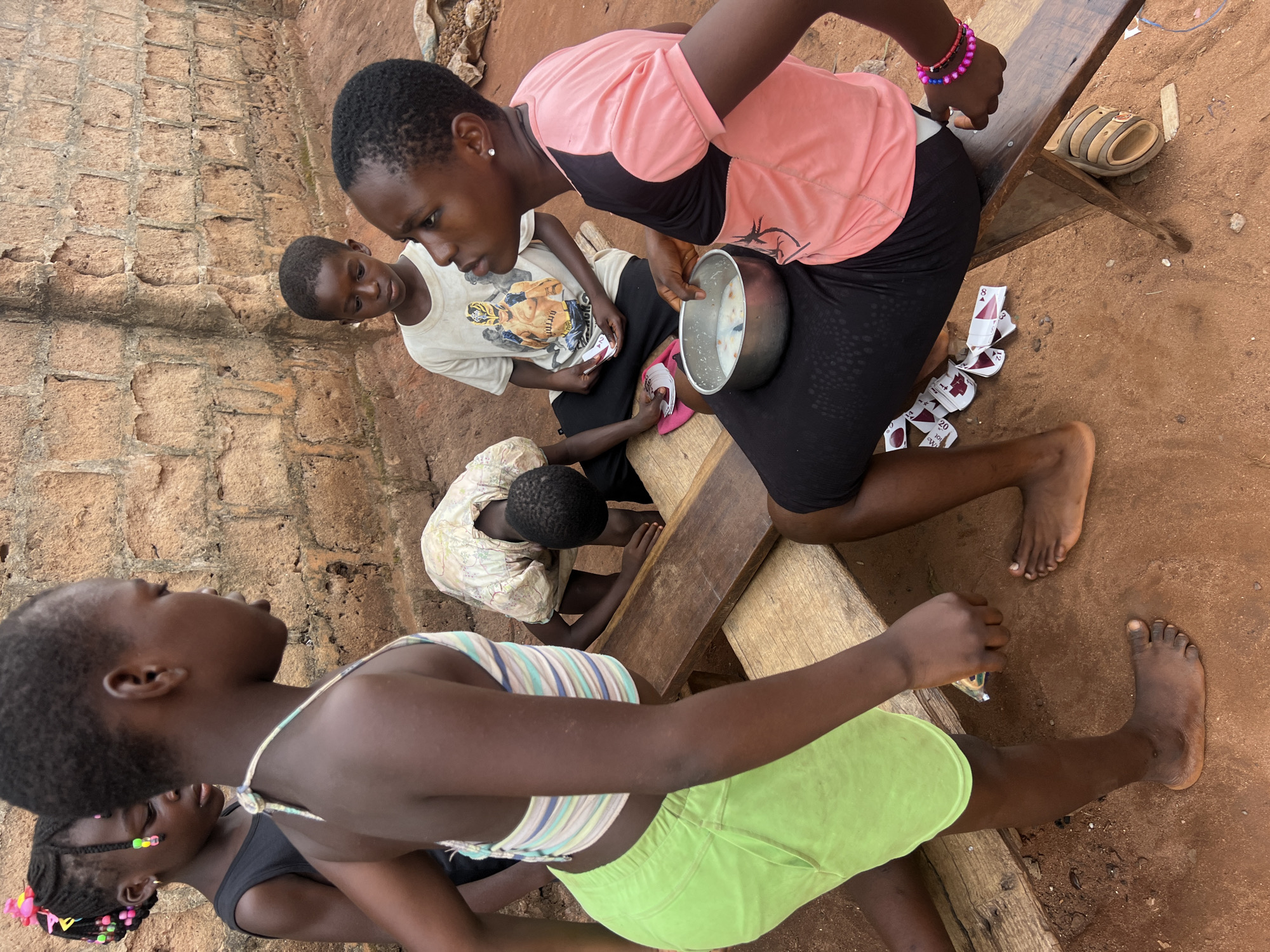 In Obosi, Nigeria, a group of children are gathered outdoors on a red-brown dirt surface. A brick wall serves as a backdrop. One child wears a light green outfit and stands in the foreground. Another child, wearing a pink top and black skirt, sits on a wooden bench, holding a metal bowl. Playing cards are scattered on the ground near her. Other children stand or sit nearby. One wears a t-shirt with a graphic design.
