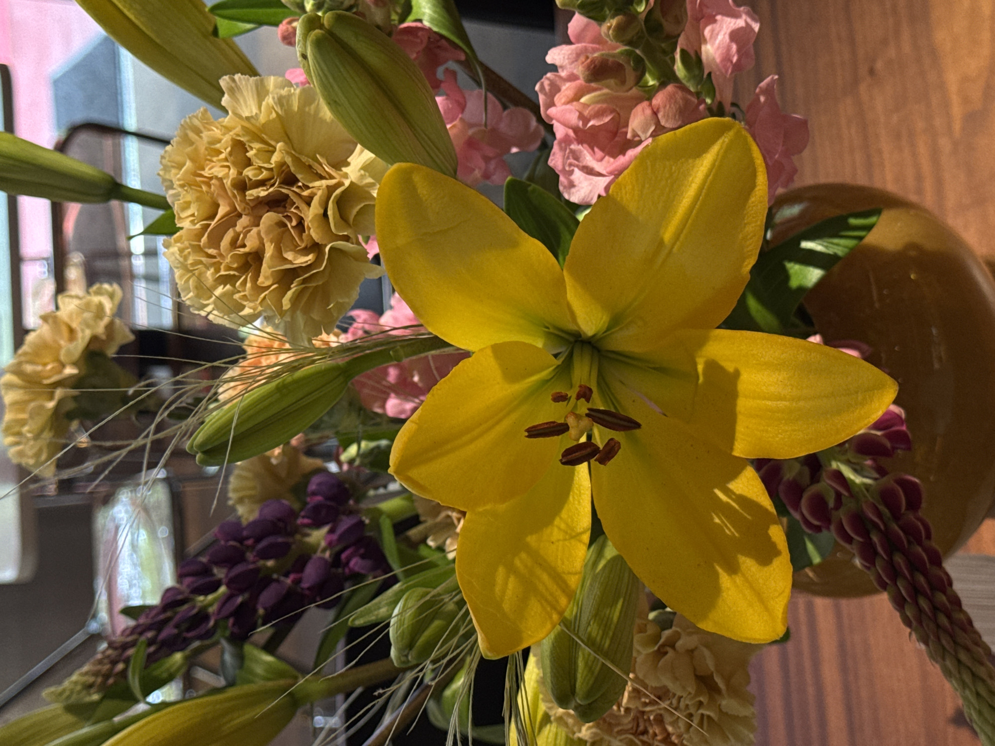 The image depicts a vibrant bouquet of flowers. Dominating the composition is a bright yellow lily with six petals and dark brown stamens. Around the lily are other flowers, including tan carnations and pink stock flowers. There are also some purple lupine-like flowers. Green lily buds and delicate blades of grass add texture to the arrangement. The bouquet is in a vase, with a light brown, possibly wooden, background visible behind the flowers. The scene seems to be indoors, with muted daylight illuminating the floral arrangement. The specific location in Yerevan, Armenia, cannot be determined from the image alone.