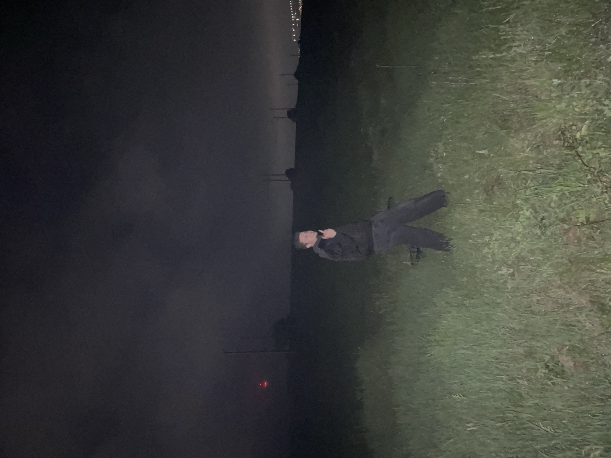A man wearing dark clothing is lying on green grass. The left side of the image is black, suggesting darkness or a shadowed area. In the distance, lights are visible, possibly from a city. A red light is visible in the darkened area. The location is likely Razgrad, Bulgaria.